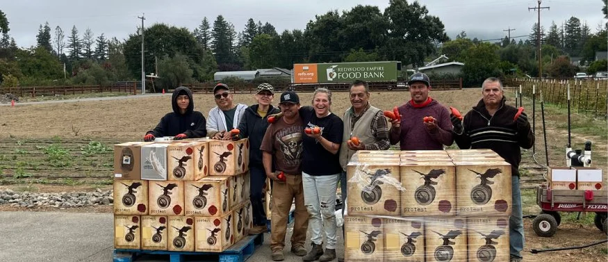 Group of seven people standing outdoors, holding tomatoes, with boxes of produce, in front of a farm field and a food bank sign.