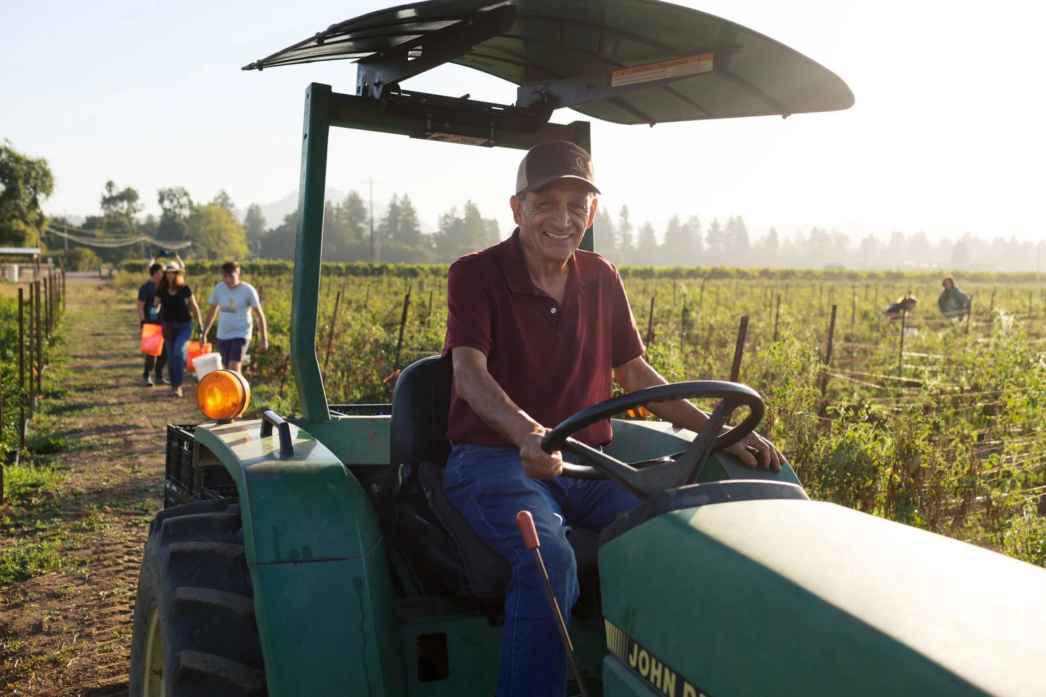 A man smiling while sitting on a green John Deere tractor in a vineyard during late afternoon. In the background, three people carrying orange buckets walk along a dirt path between rows of grapevines, with trees and a clear sky in the distance.