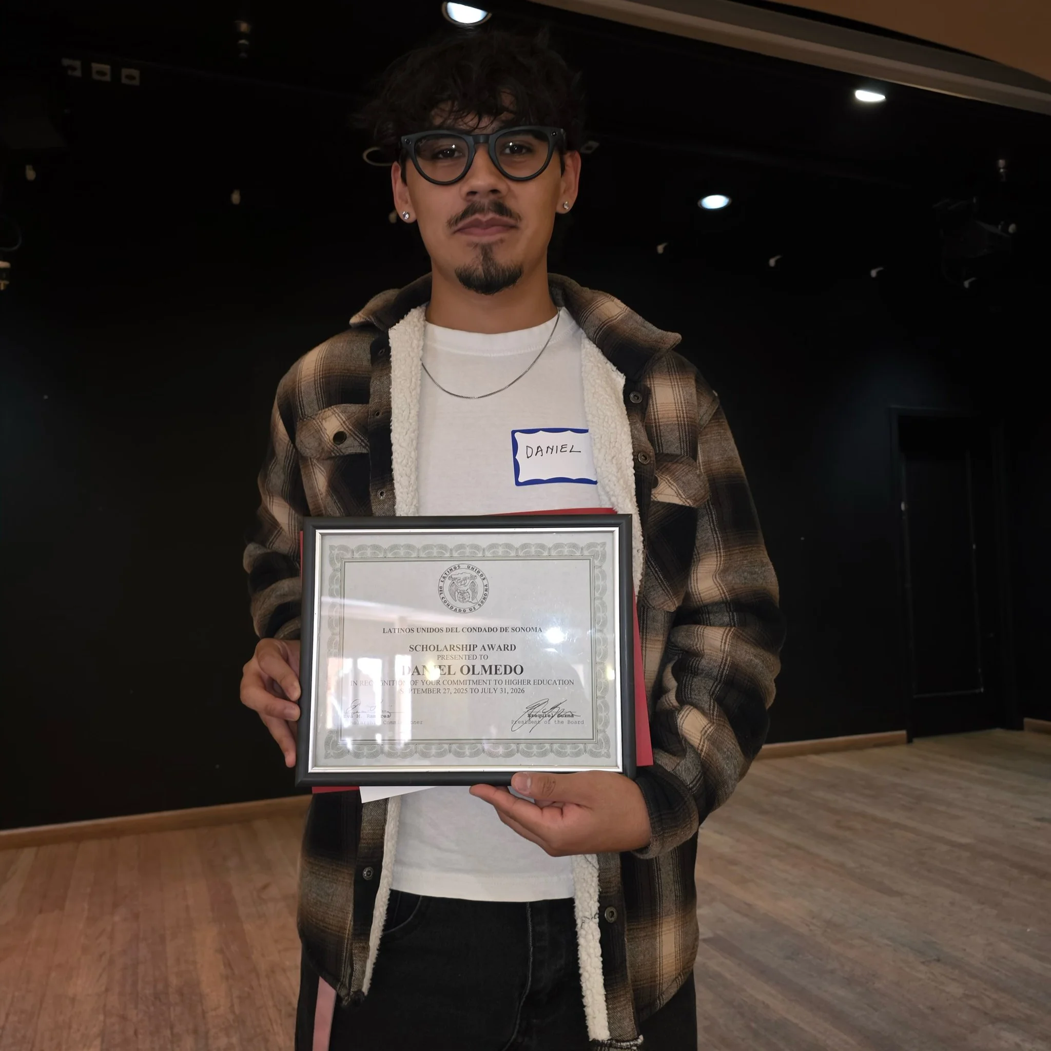 A young man with dark, curly hair and glasses holding a certificate of scholarship award at an indoor event. He is wearing a white shirt, a plaid jacket with sherpa lining, and has a name tag that says Daniel.