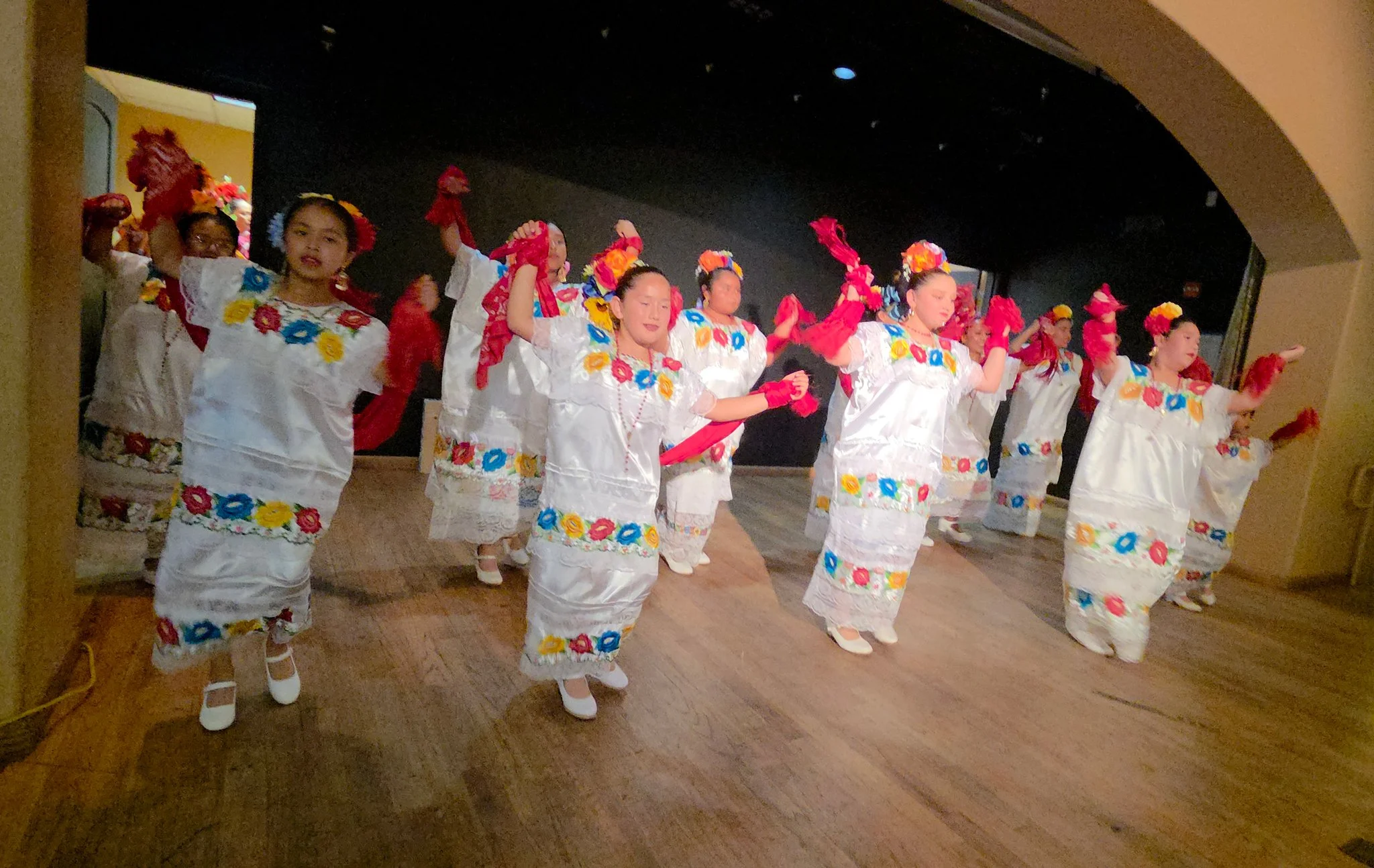 Group of women dressed in white traditional dresses with colorful floral embroidery, performing a dance on stage, some with red accessories and floral headpieces.