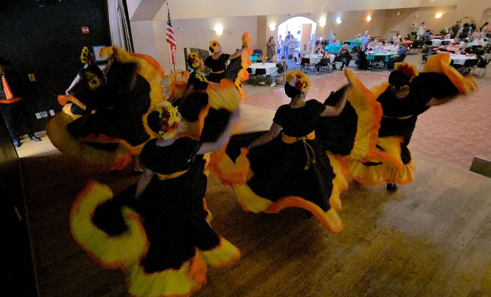 People dressed in colorful costumes perform a dance in a decorated hall with tables and people seated.