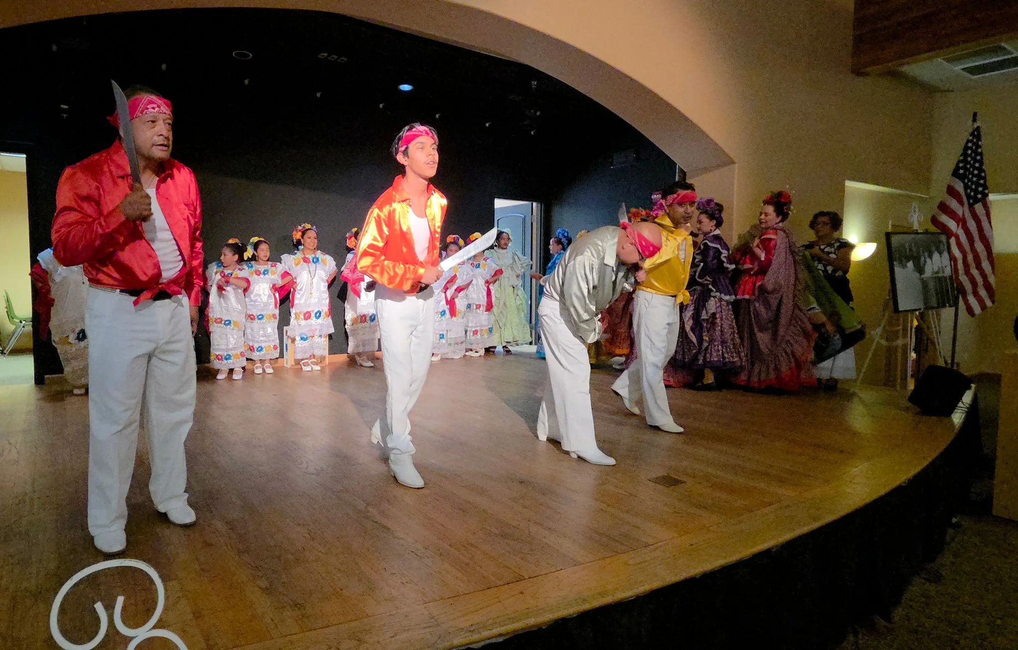 Group of people performing in a cultural dance on stage, dressed in traditional Mexican attire, with some women in colorful dresses and men in white pants and colorful shirts, and a Mexican flag in the background.