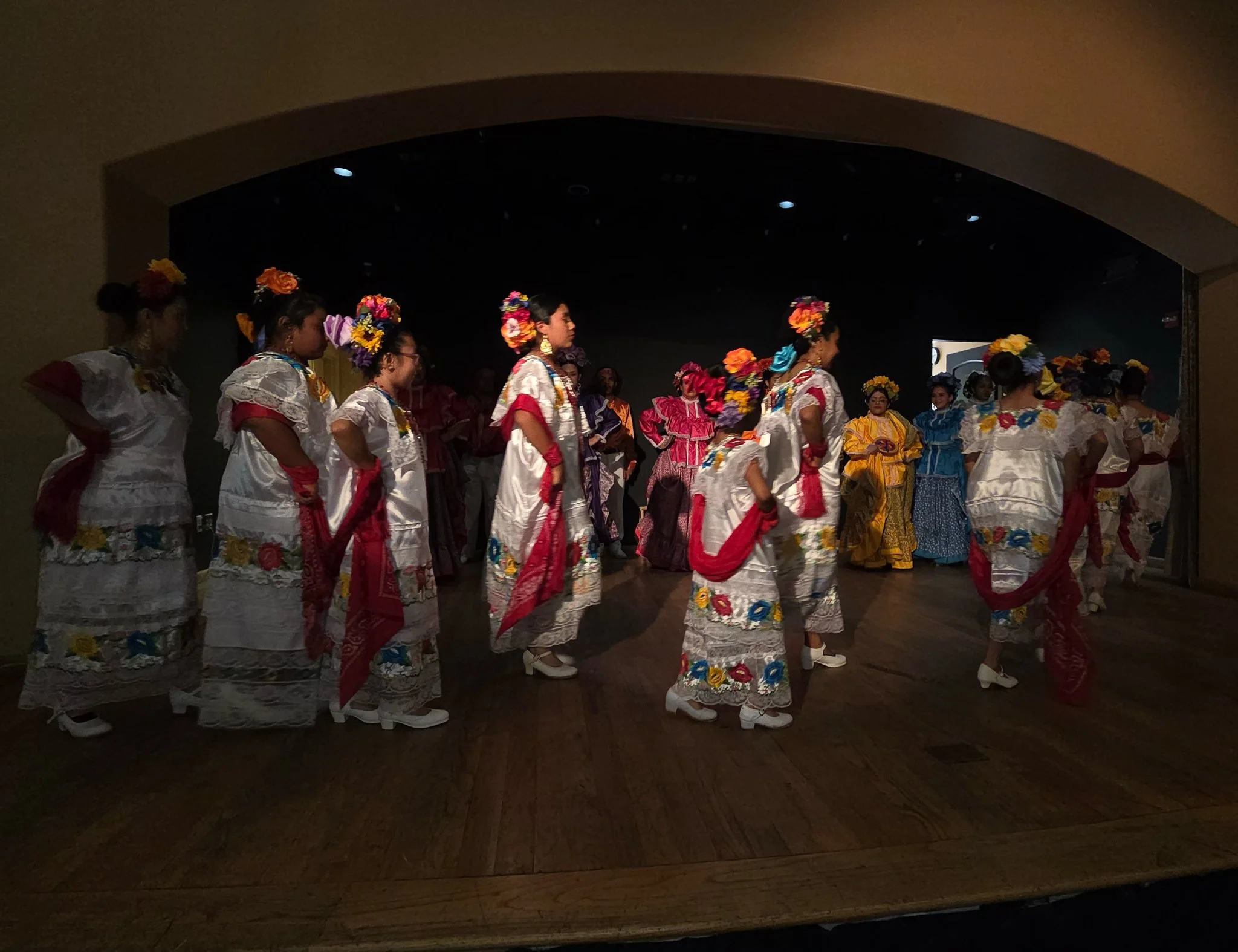 Group of women dressed in colorful traditional folkloric dresses participating in a dance performance on stage.