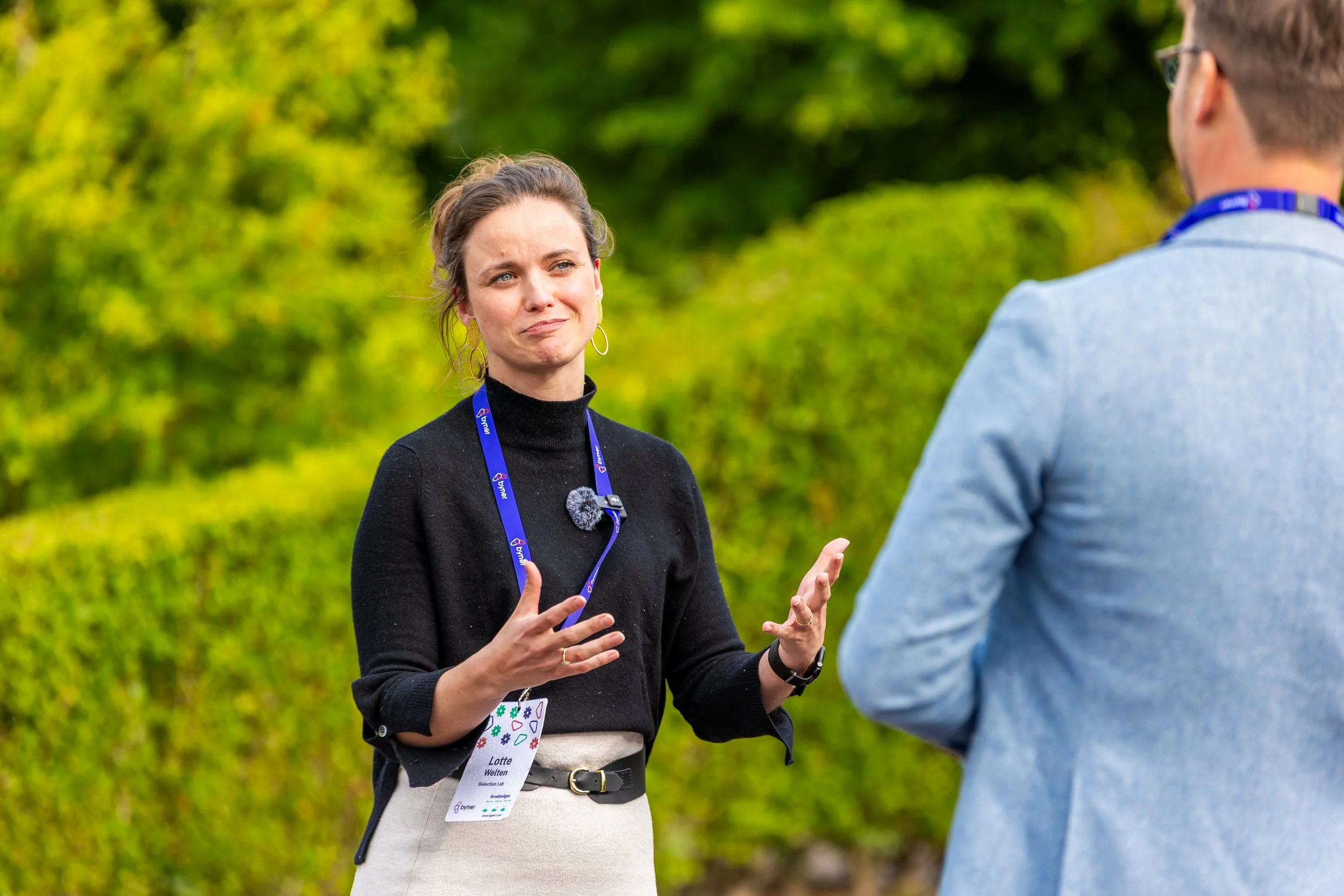 Een vrouw die in gesprek is met een man buiten, met groene struiken op de achtergrond.