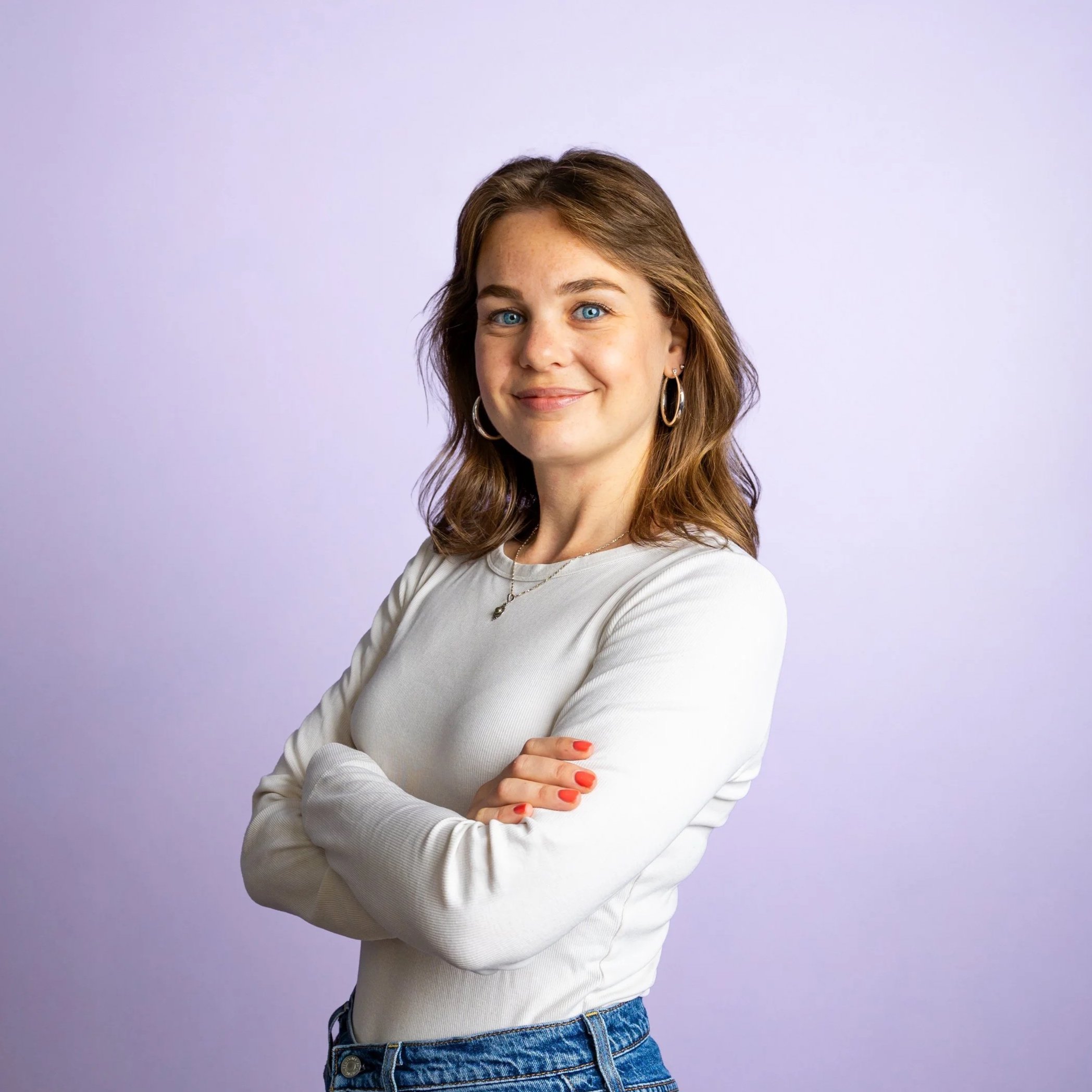 Een jonge vrouw met blauwe ogen, bruin haar en lichte huid, gekleed in een wit topje en blauwe jeans, poseert met haar armen over elkaar tegen een lichte paarse achtergrond.