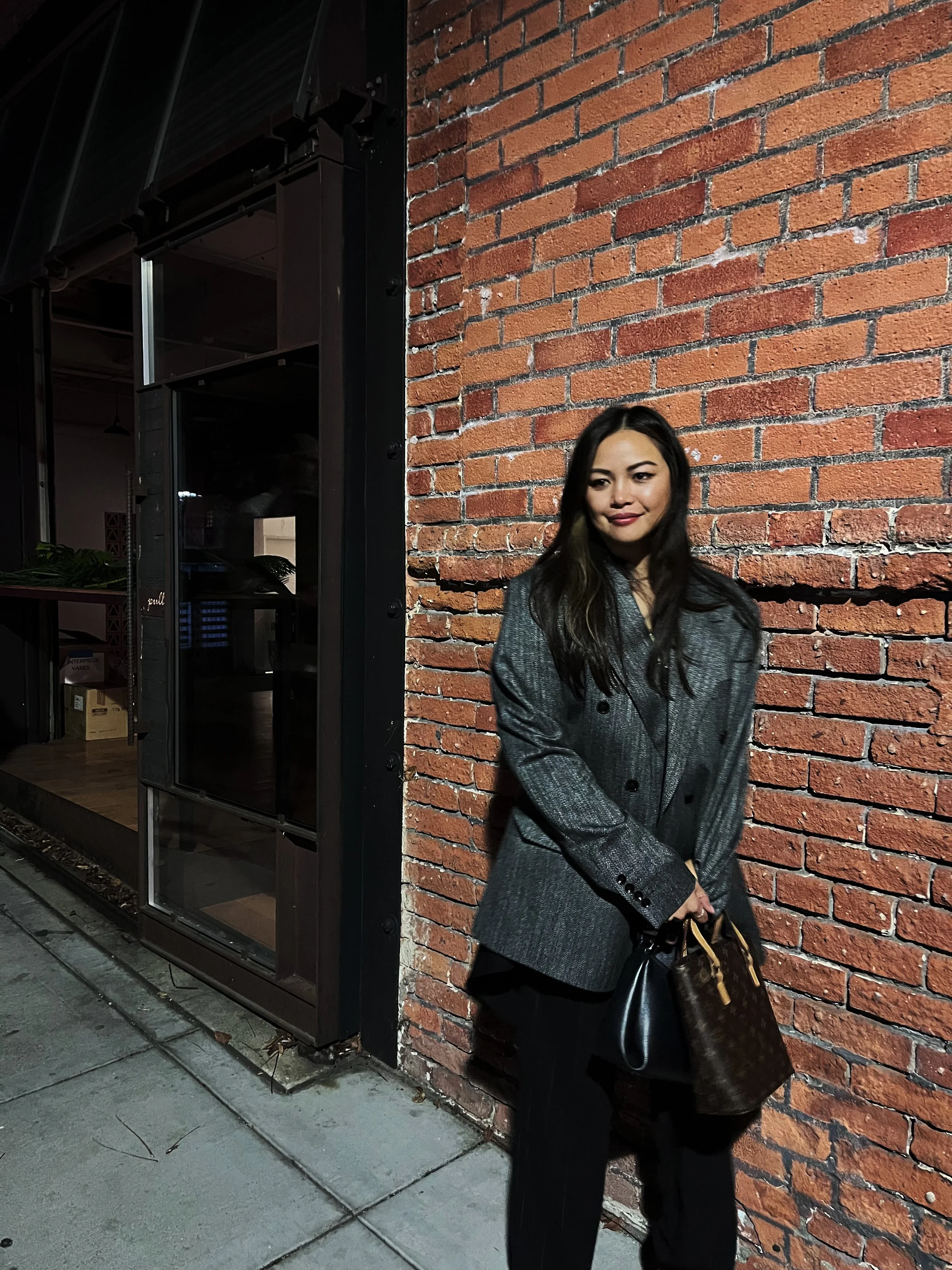 A woman stands outside against a brick wall at night, holding handbags, wearing a gray blazer, with dark hair and makeup.