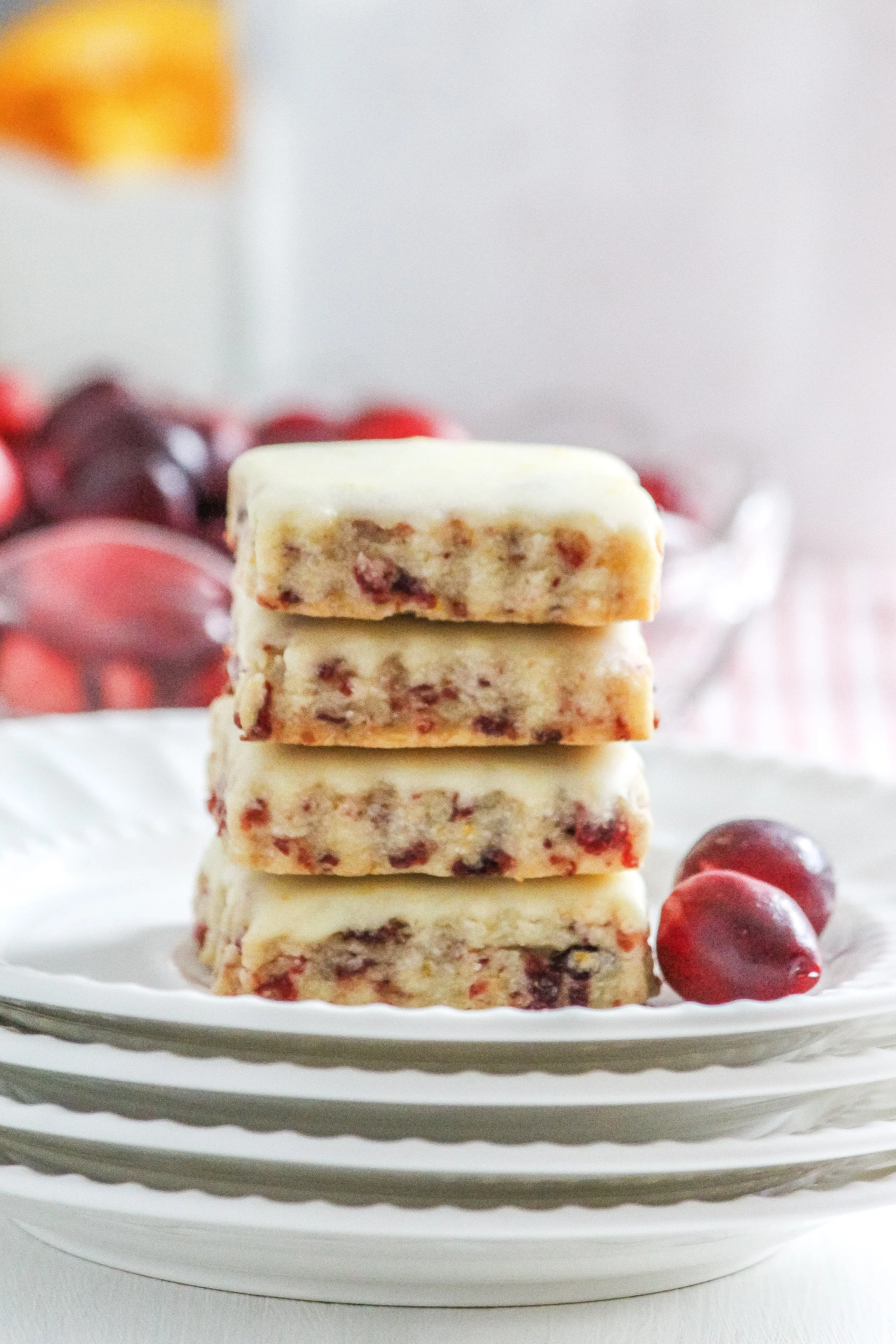 Stack of four white and red cherry fudge pieces on a white plate with a few cherries beside them, with a blurred bowl of cherries in the background.