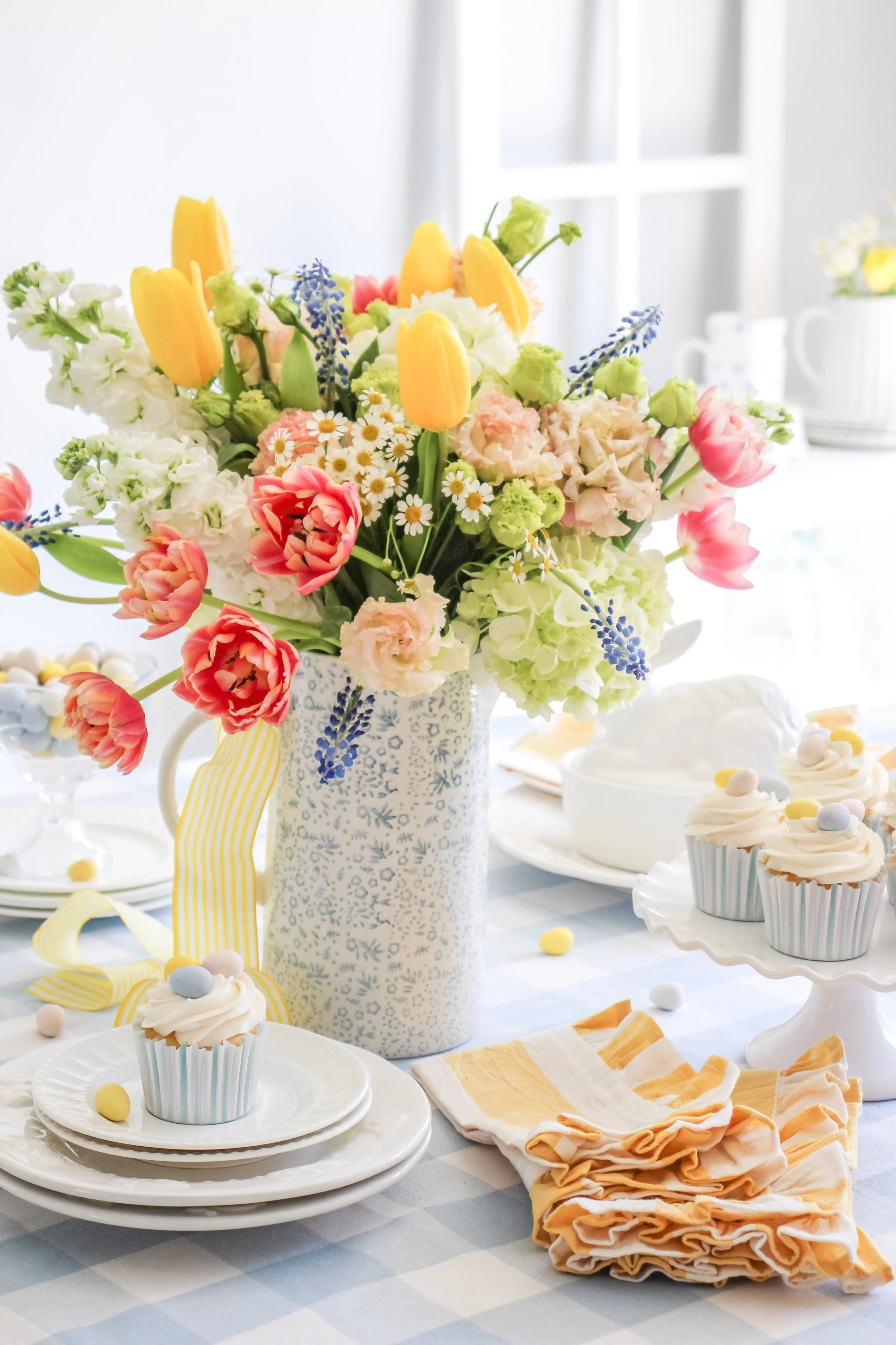 Colorful spring table setting with a large floral bouquet of tulips, daisies, and other flowers in a patterned white vase, cupcakes with pastel sprinkles, and a cake on a white cake stand, all on a blue and white checkered tablecloth.