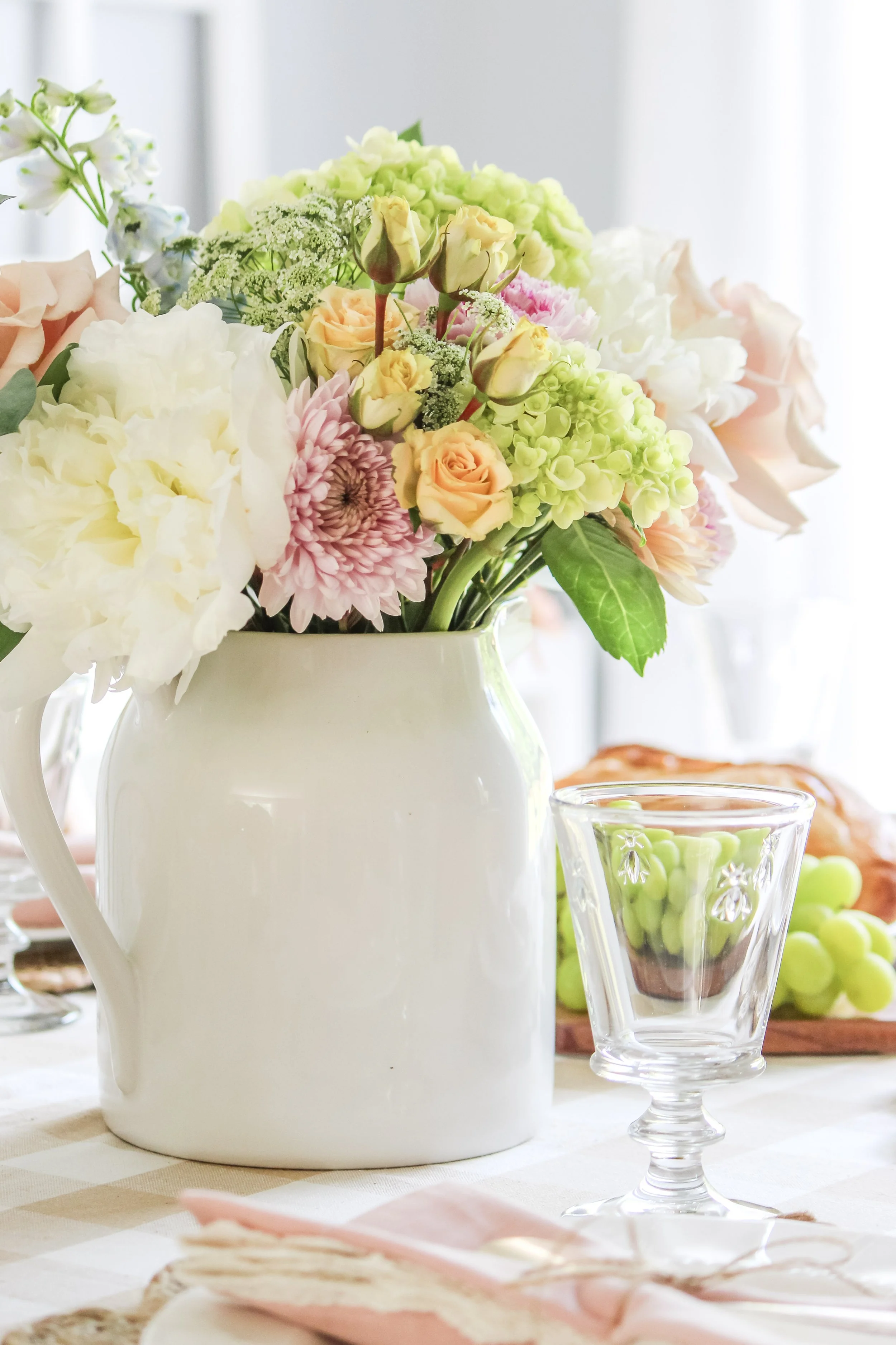 A white ceramic pitcher filled with a colorful bouquet of flowers, including roses and hydrangeas, on a table with a glass container holding green grapes.