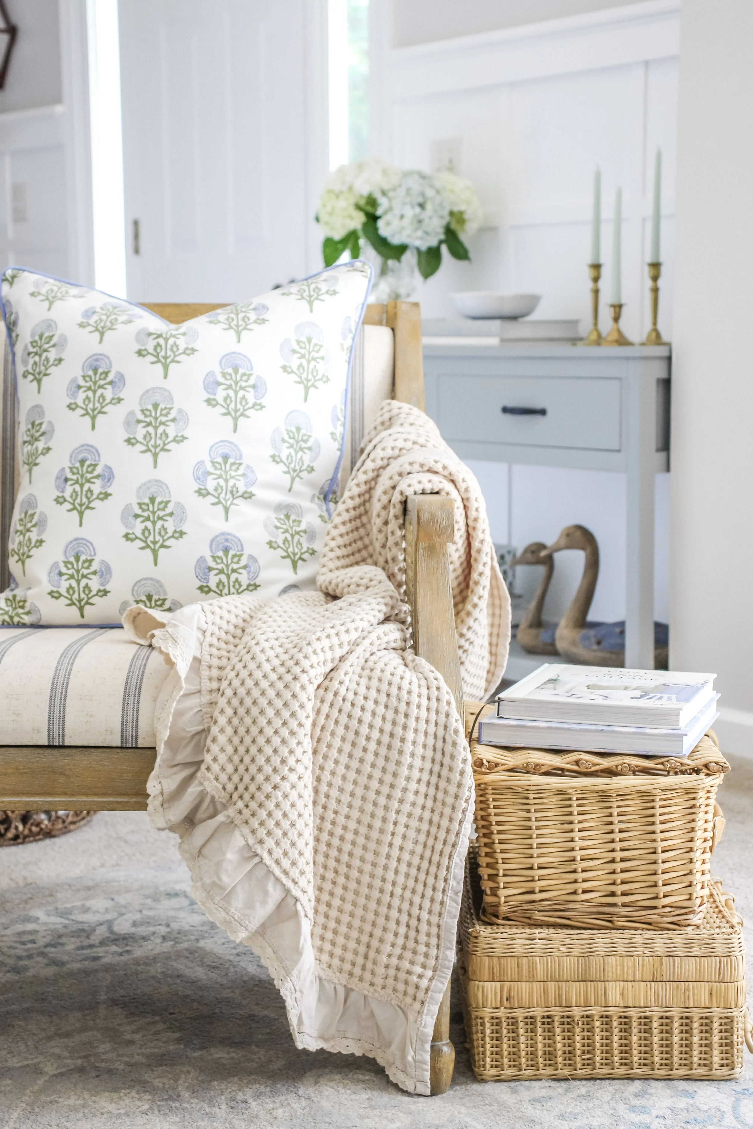 A cozy indoor scene with a cushioned wooden bench, decorative pillows, a blanket, a wicker side table with magazines, a sideboard with a floral bouquet, candleholders, and decorative swan figurines.