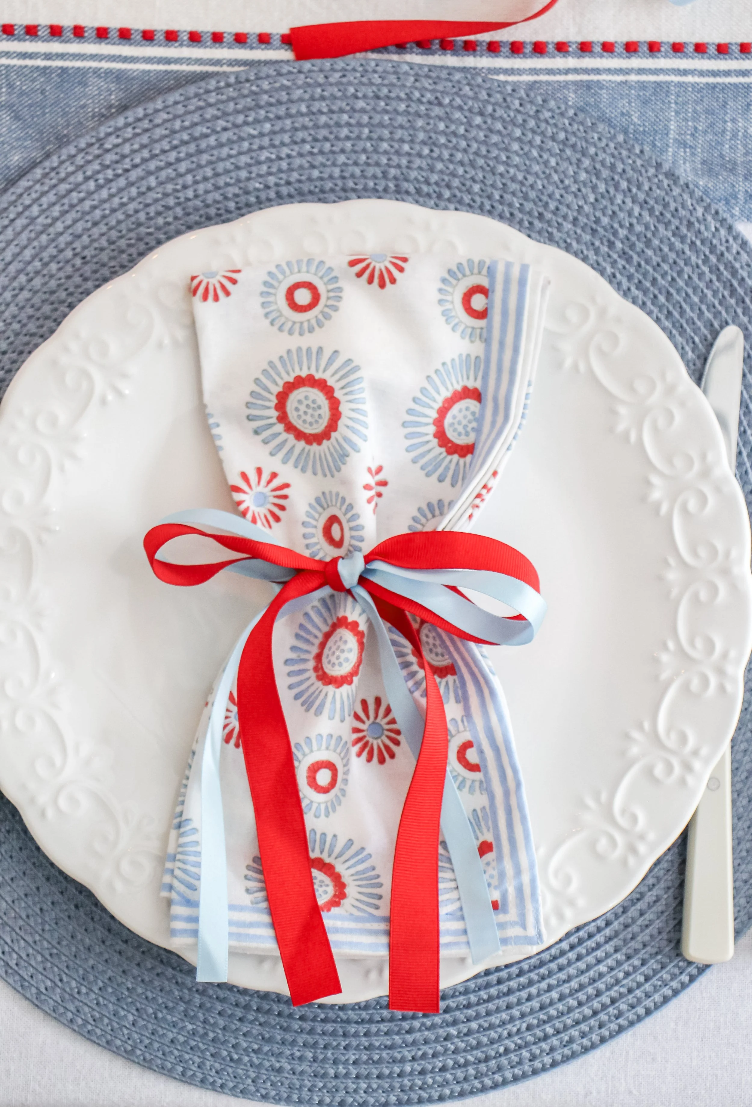 A white ceramic plate with an ornate border, set on a blue woven placemat. A folded patterned paper napkin with red, light blue, and gray floral designs is placed on the plate and tied with two ribbons, one red and one light blue.