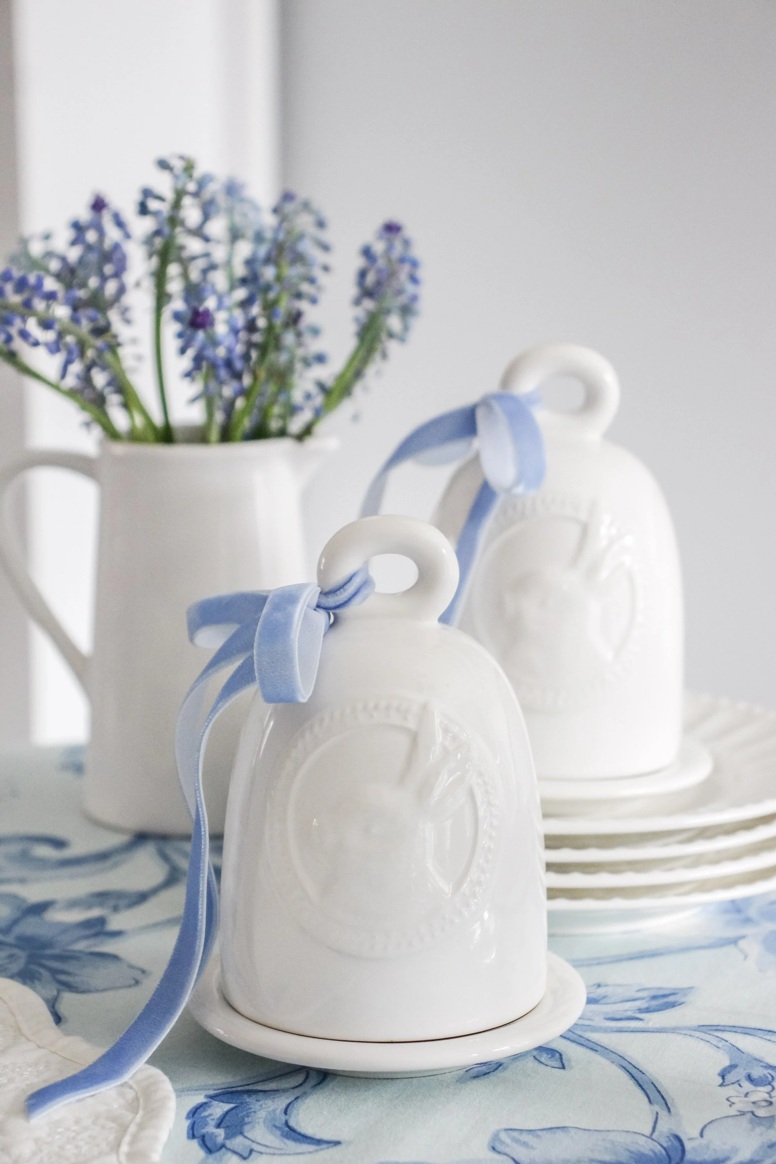 White ceramic bells with blue ribbons, stacked on saucers, on a table with a blue floral tablecloth, with a white pitcher of purple flowers in the background.