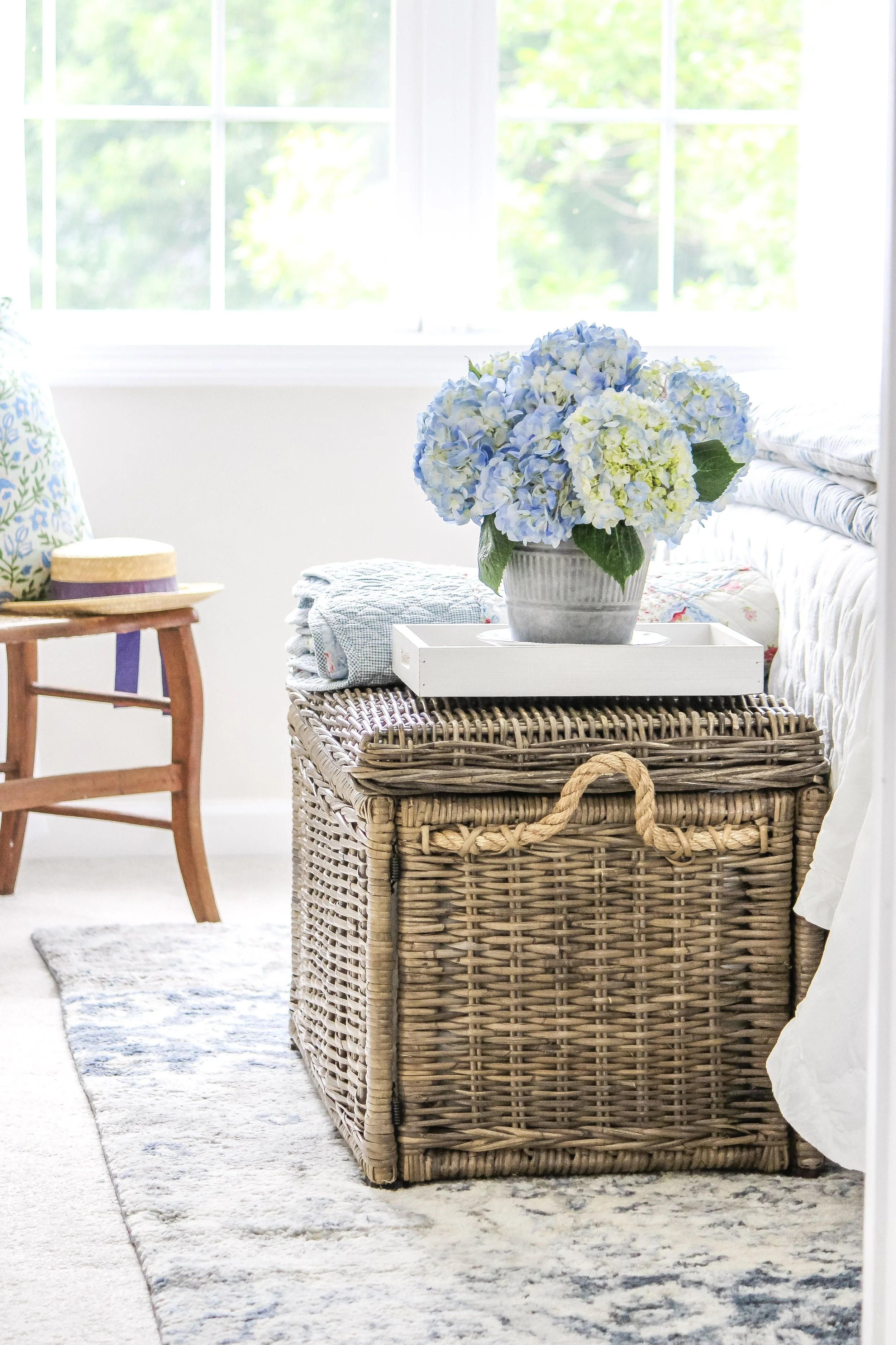 A wicker storage basket with a white tray on top, holding a vase of blue and white hydrangeas, next to a bed with a quilt, in a bright room with a window and a wooden chair.
