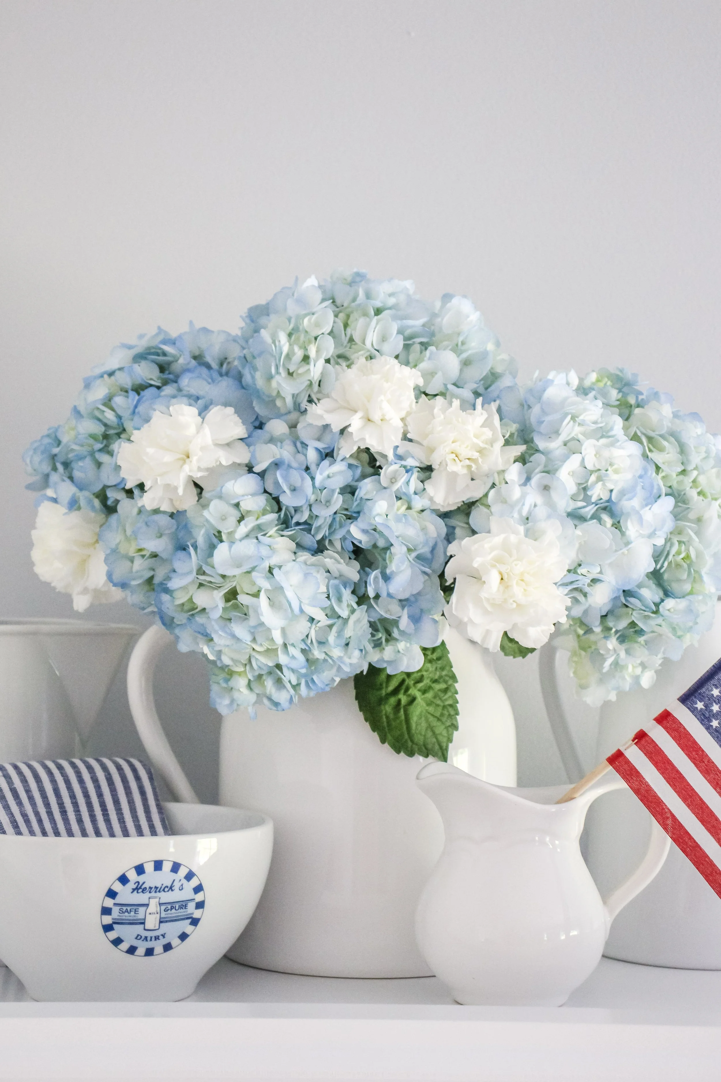 A bouquet of light blue and white hydrangeas and white carnations in a white vase, with a corner of a milk bowl with a blue striped cloth and an American flag in a white creamer jug.