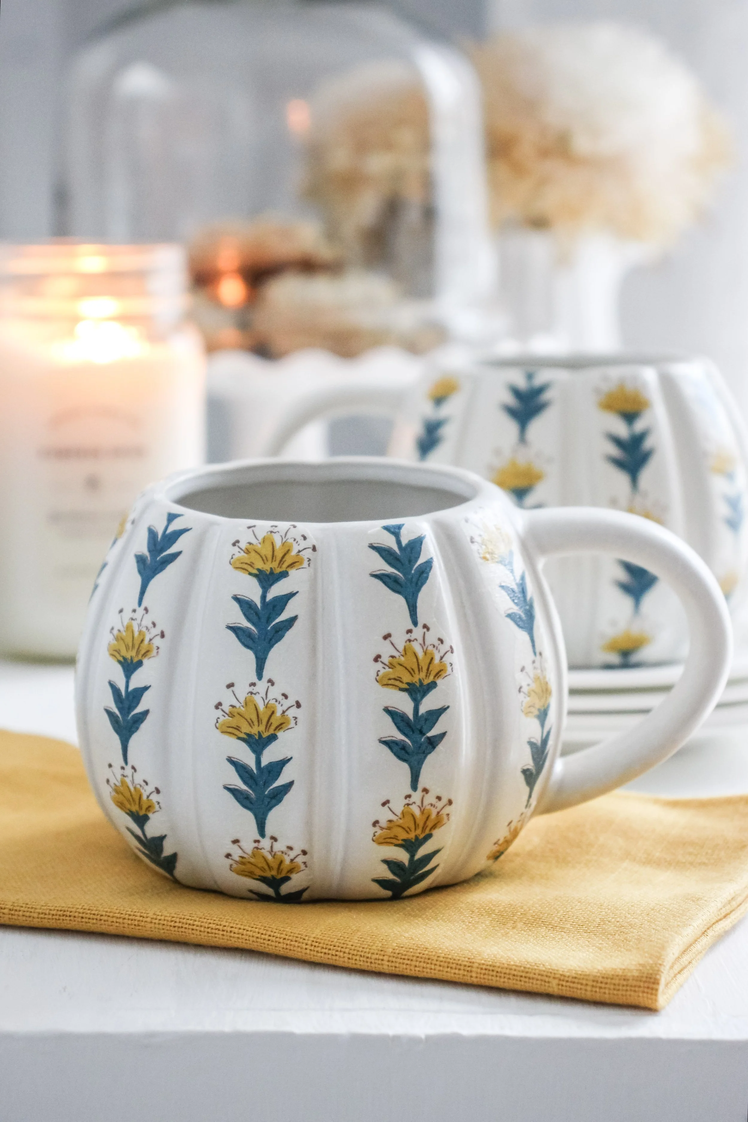 A decorative white mug with yellow and blue floral patterns sitting on a yellow cloth napkin. In the background, there is a lit candle, another similar mug, and a glass cloche covering a stack of cookies, with a blurred vase of flowers behind them.