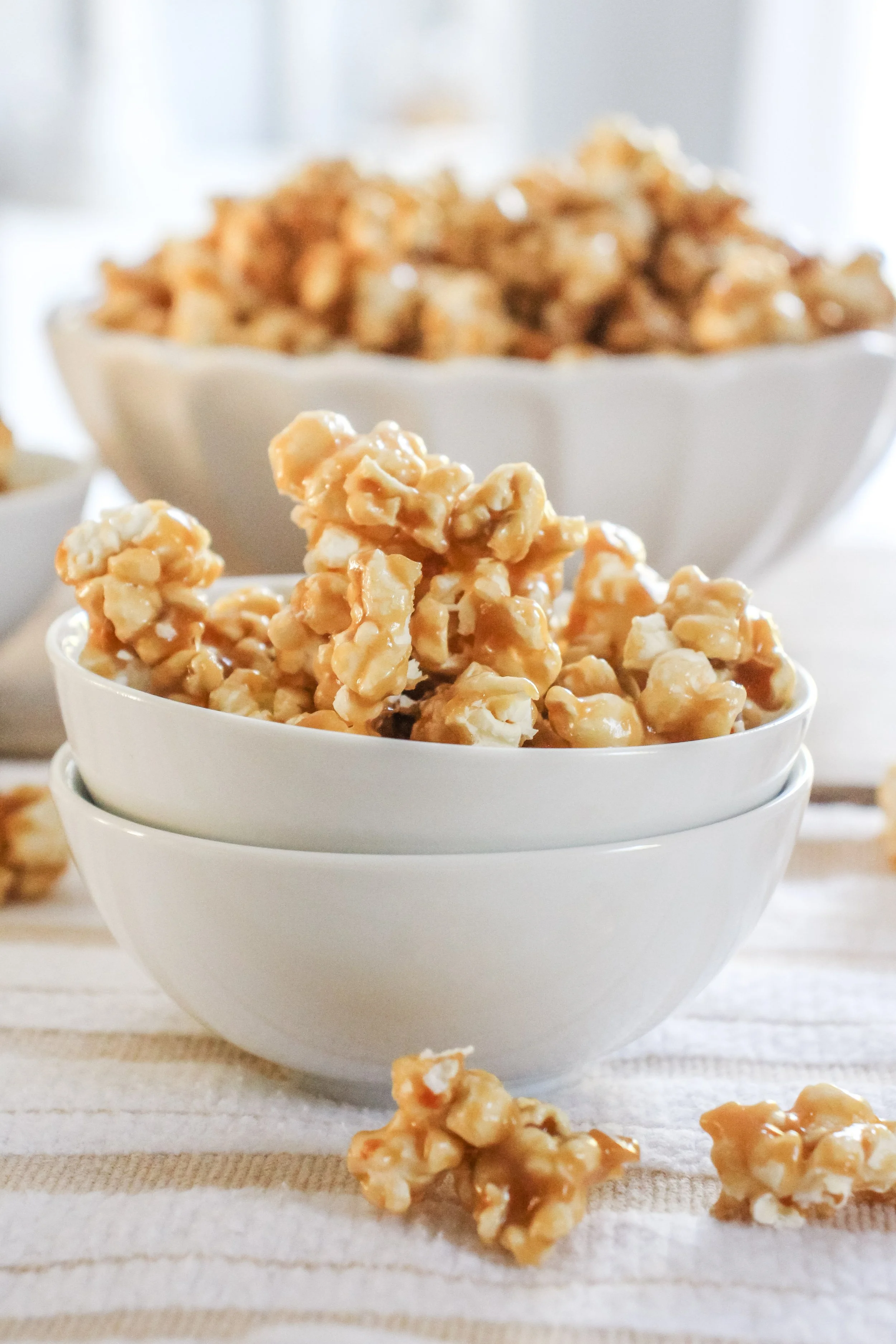 Close-up of caramel popcorn in a white ceramic bowl on a striped cloth, with additional popcorn in background.