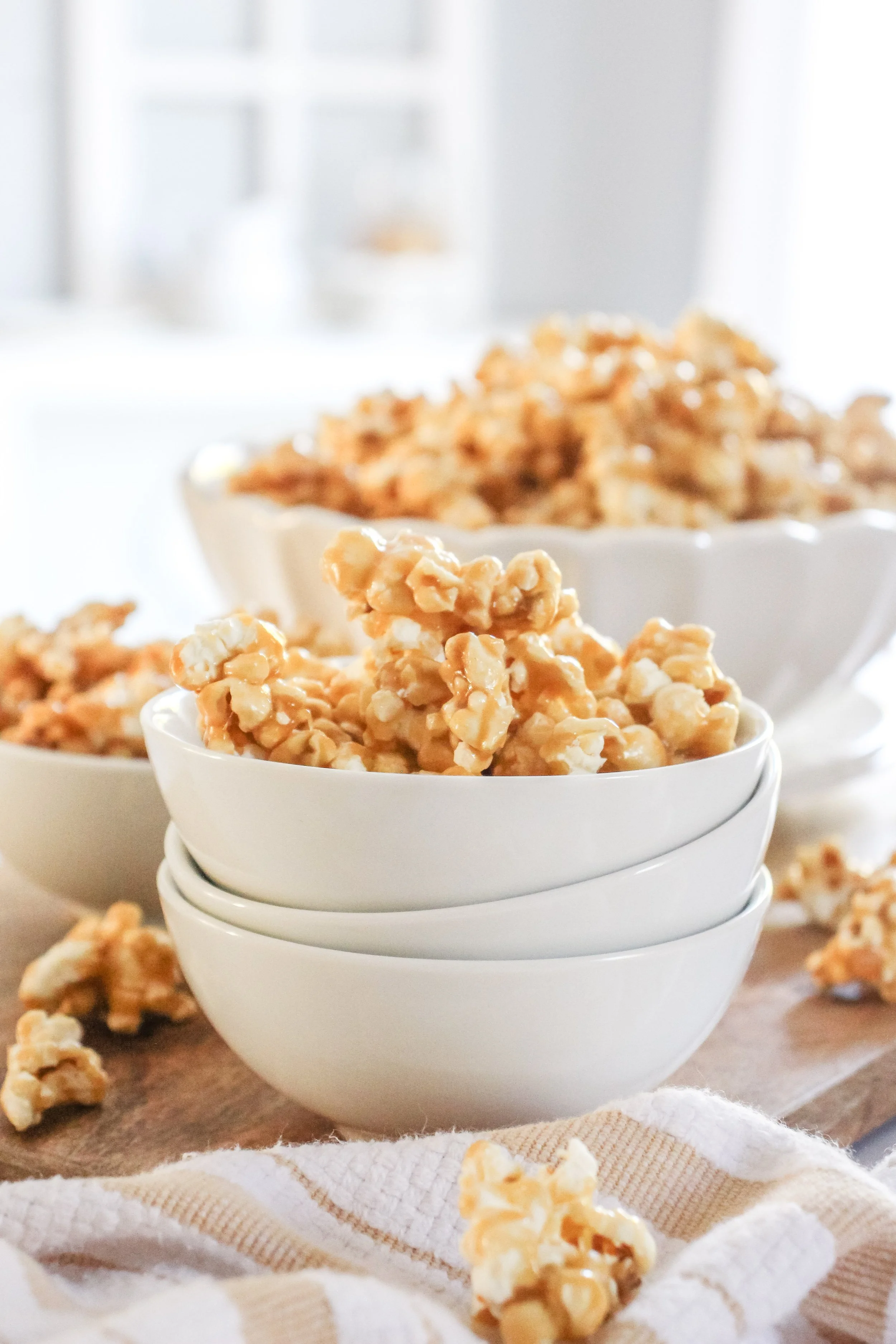 Stacked white bowls filled with caramel popcorn on a wooden surface, with additional popcorn scattered around.