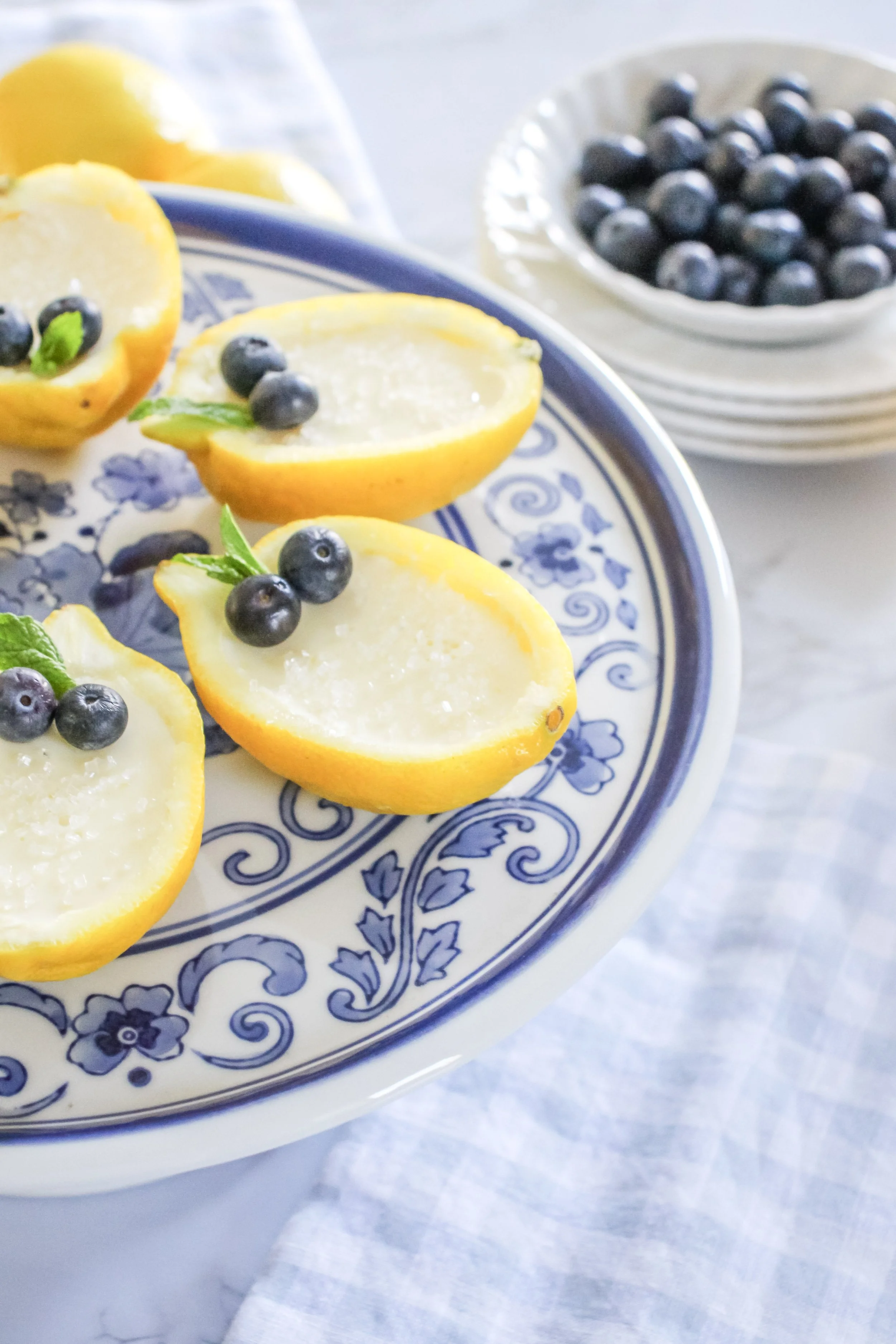 Halved lemons filled with a creamy mixture, garnished with blueberries and mint leaves on a decorative blue and white plate, with a bowl of blueberries in the background.