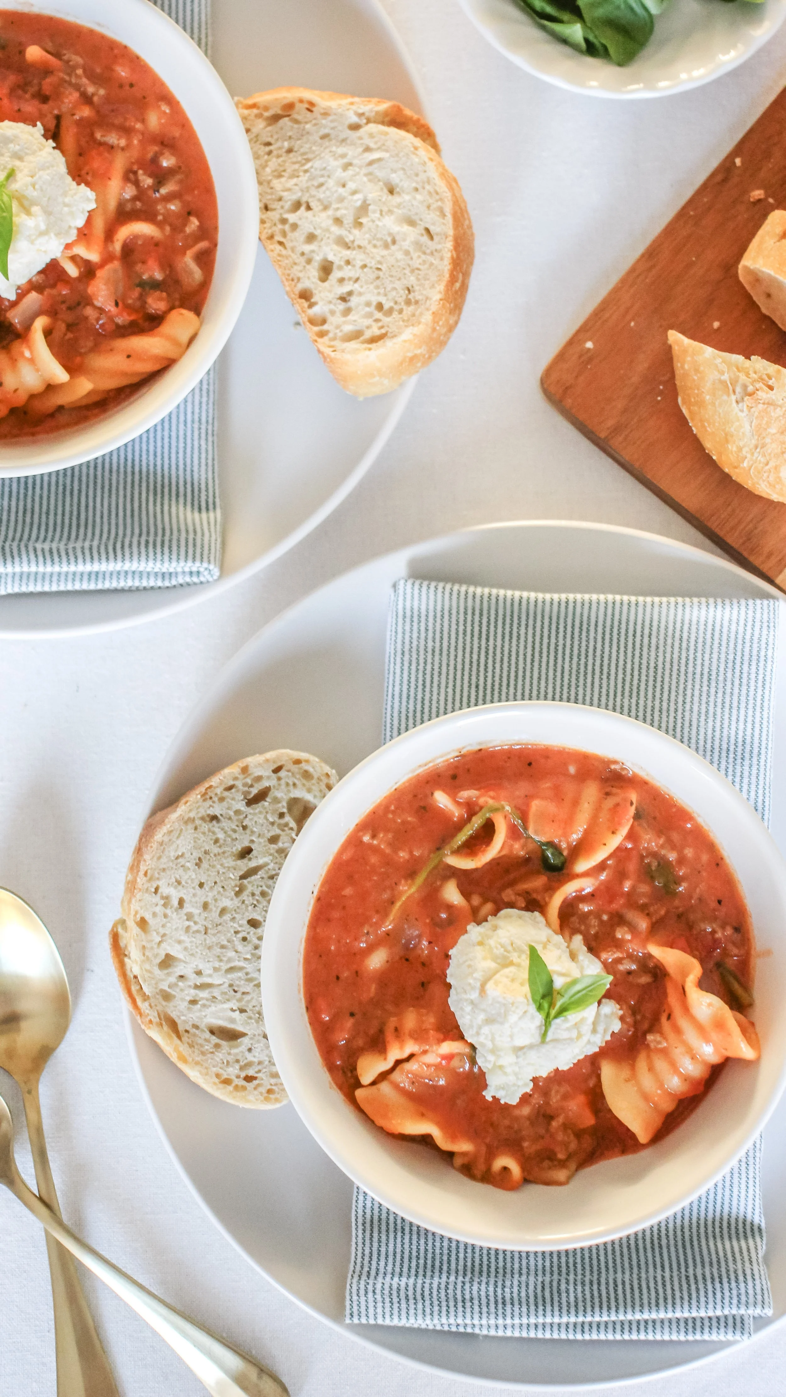 Two bowls of pasta soup with a dollop of sour cream and basil on top, served with slices of bread on a white plate, napkins, and gold-colored spoons on a white table.