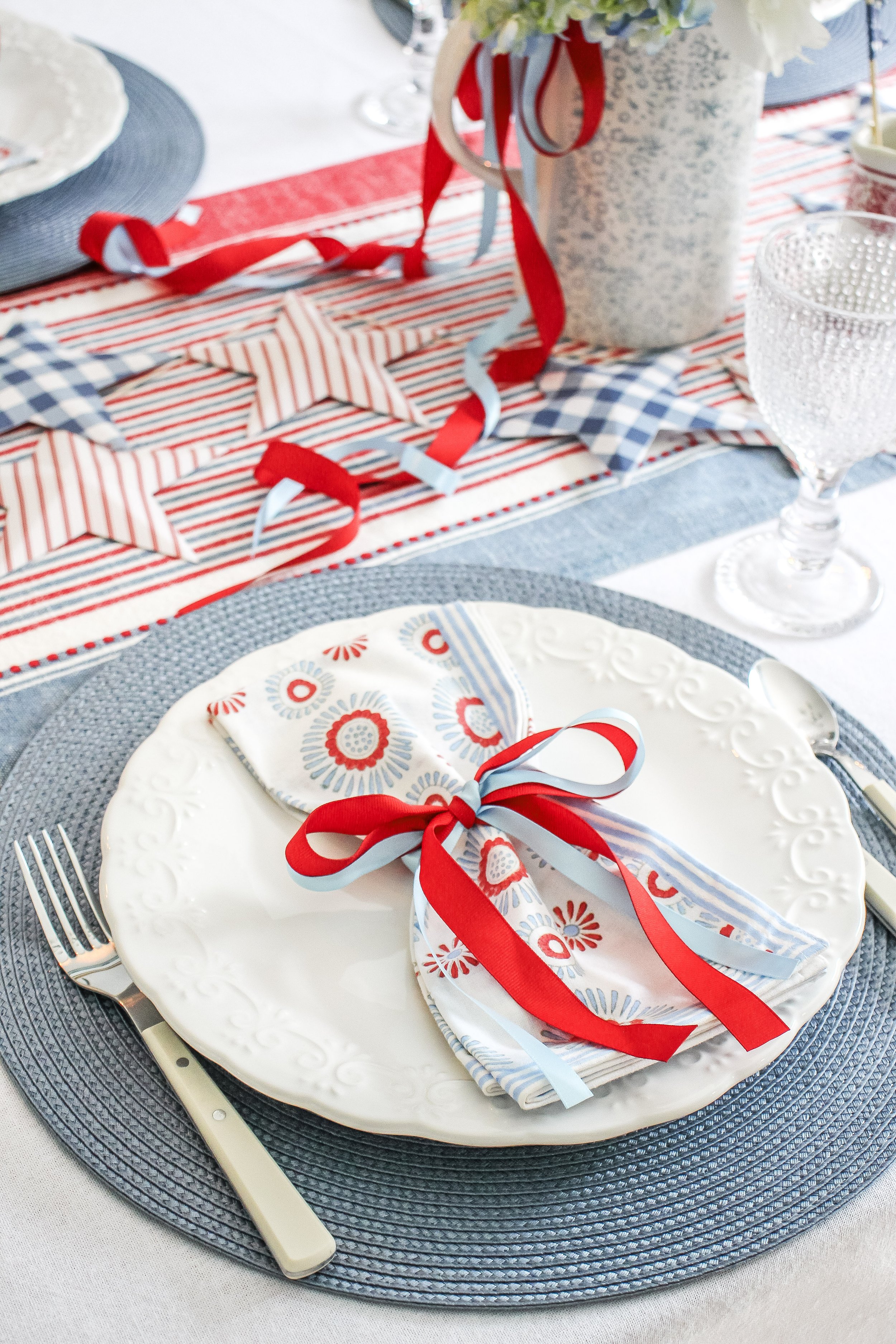 A decorated table with red, white, and blue patriotic theme, featuring a white plate with a floral napkin tied with red and light blue ribbons, a fork on the left, and a glass on the right, with patriotic star and stripe decorations and flower arrang