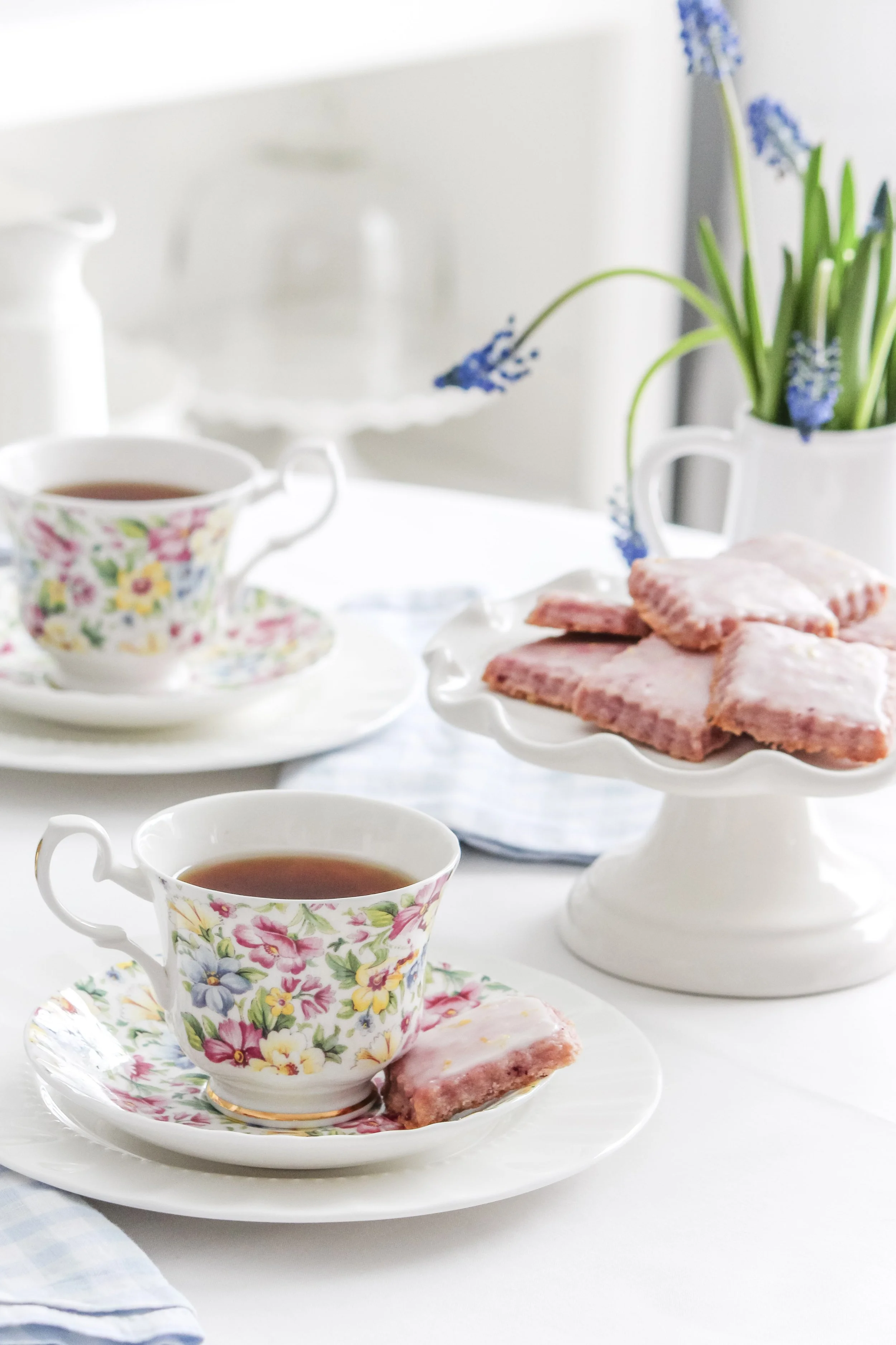 Tea cups with floral patterns, a plate with pink frosted cookies on a cake stand, a vase with blue flowers, on a white table.