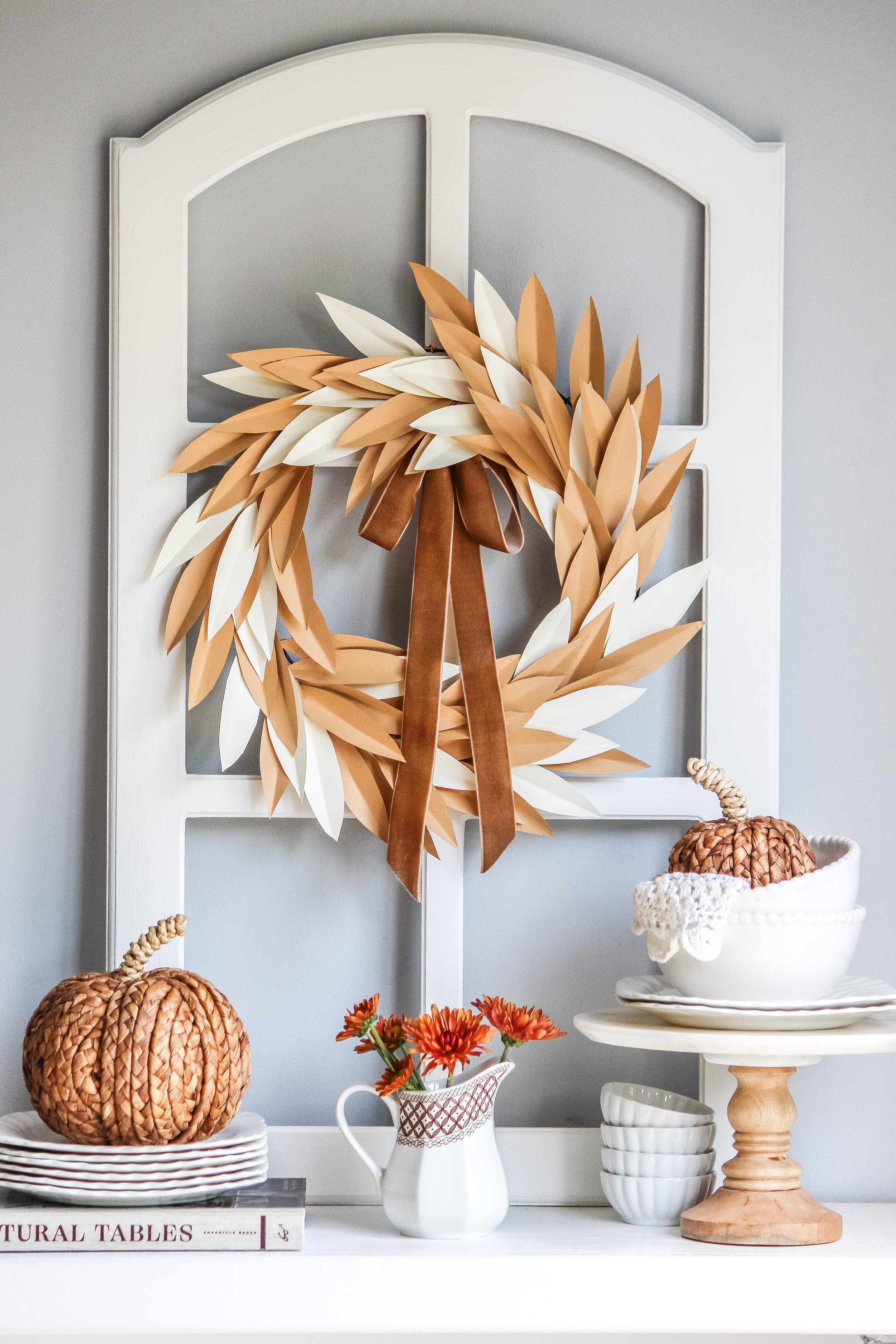 Decorative fall-themed display with a paper leaf wreath on a white window frame, along with a ceramic pumpkin, a small pitcher with orange flowers, and a two-tiered cake stand with a ceramic pumpkin and a lace doily.
