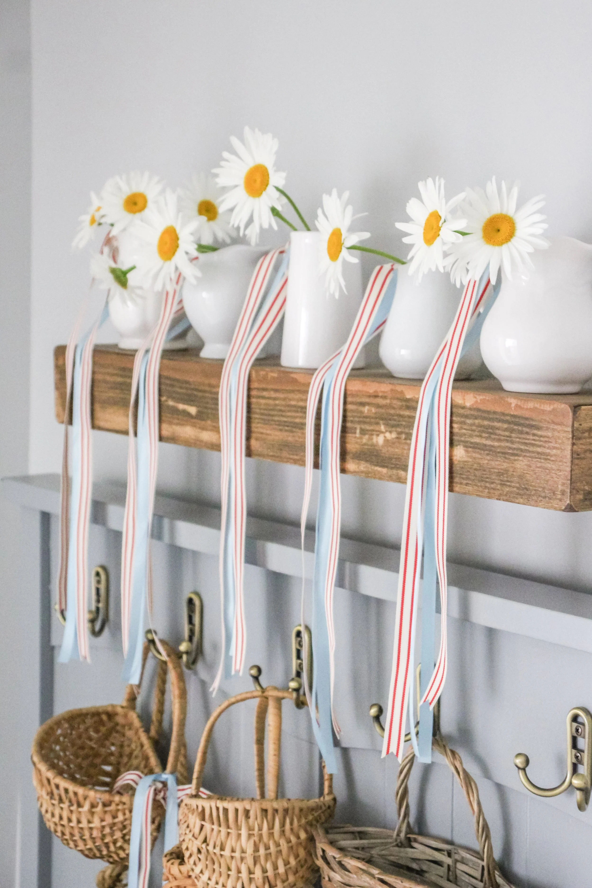 White vases with daisies and striped ribbons on a wooden shelf, below hanging baskets and hooks.