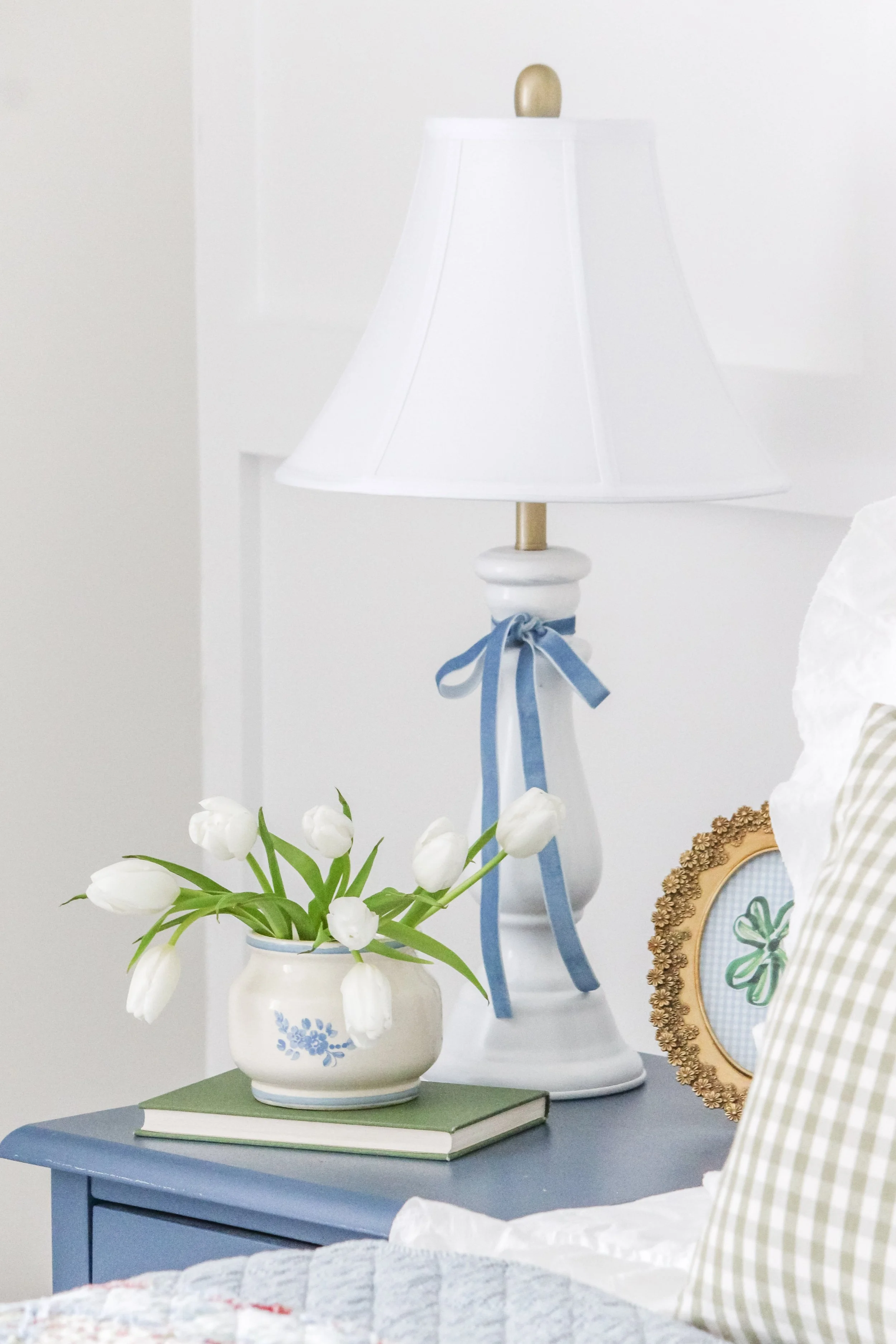 A blue bedside table with a vase of white tulips, a green book, a framed shamrock picture, and a white lamp with a blue ribbon tied around the base, in a bright bedroom.