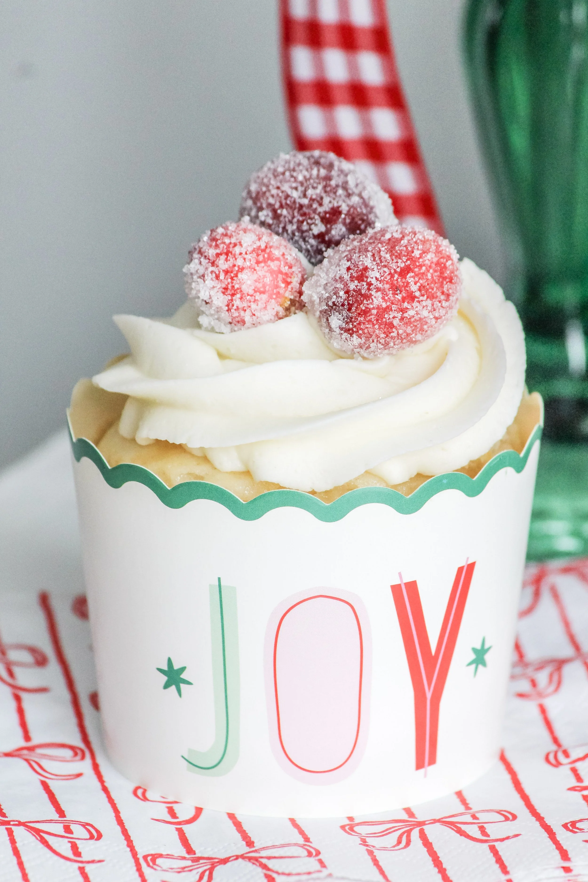 Close-up of a Christmas-themed cupcake with white frosting and sugared cherries on top, in a cup that says 'JOY' in red and green letters.