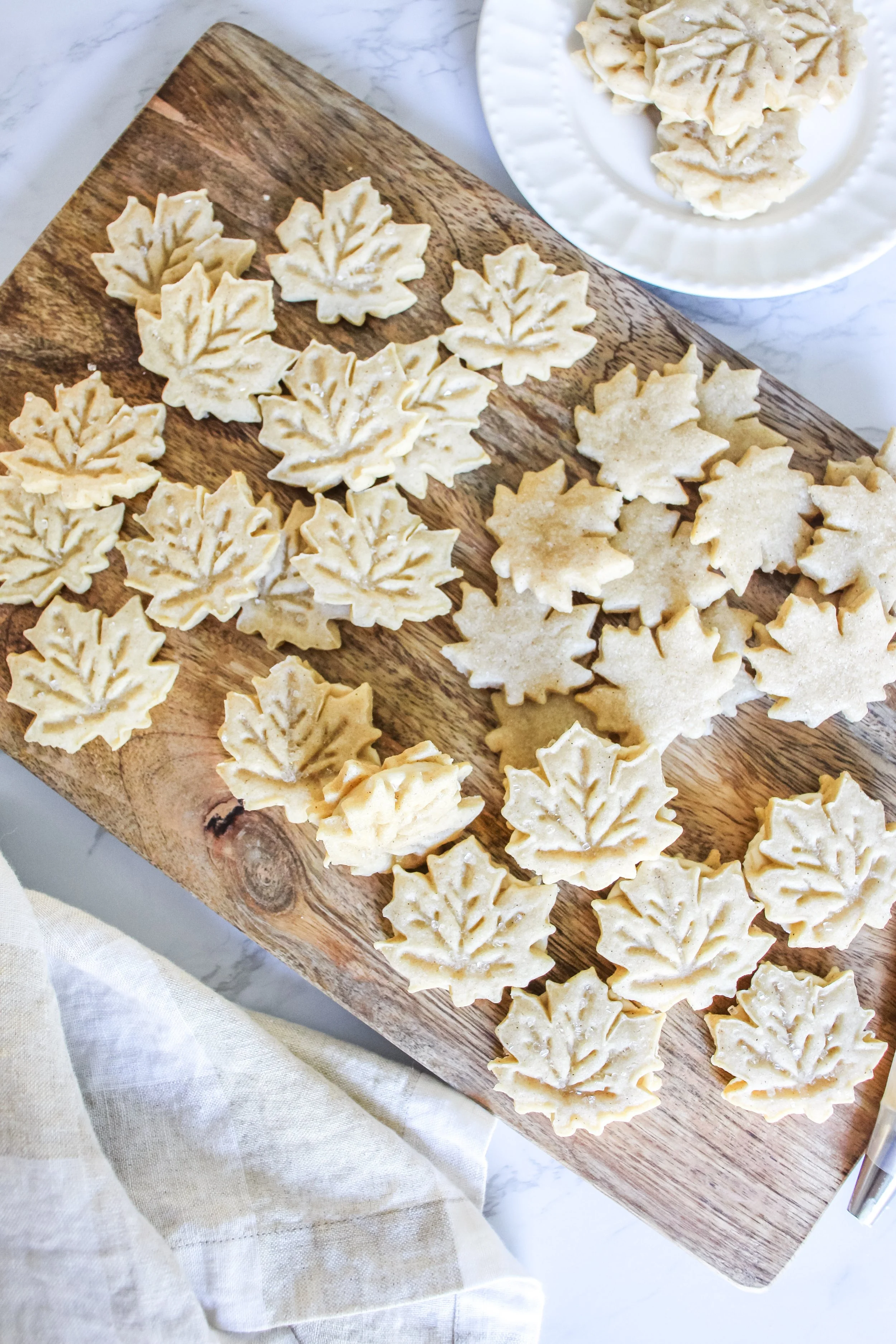 Maple leaf-shaped cookies on a wooden cutting board, with additional cookies on a white plate nearby.