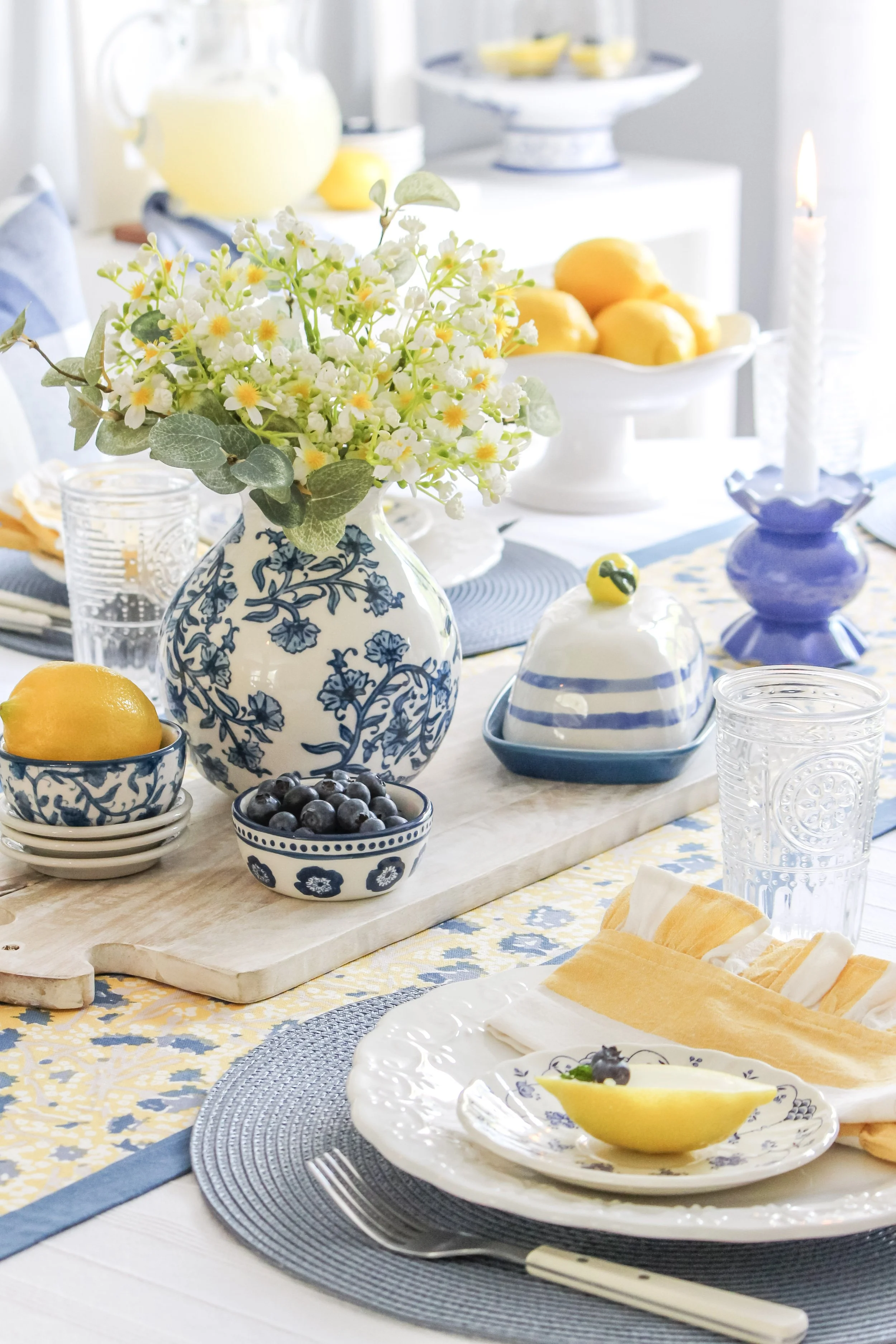 A beautifully set dining table with a blue and white color theme. There are plates, glasses, a lemon, bowl of blueberries, a floral vase, and a butter dish. Lemons are in a white bowl, and a lit candle is also on the table.
