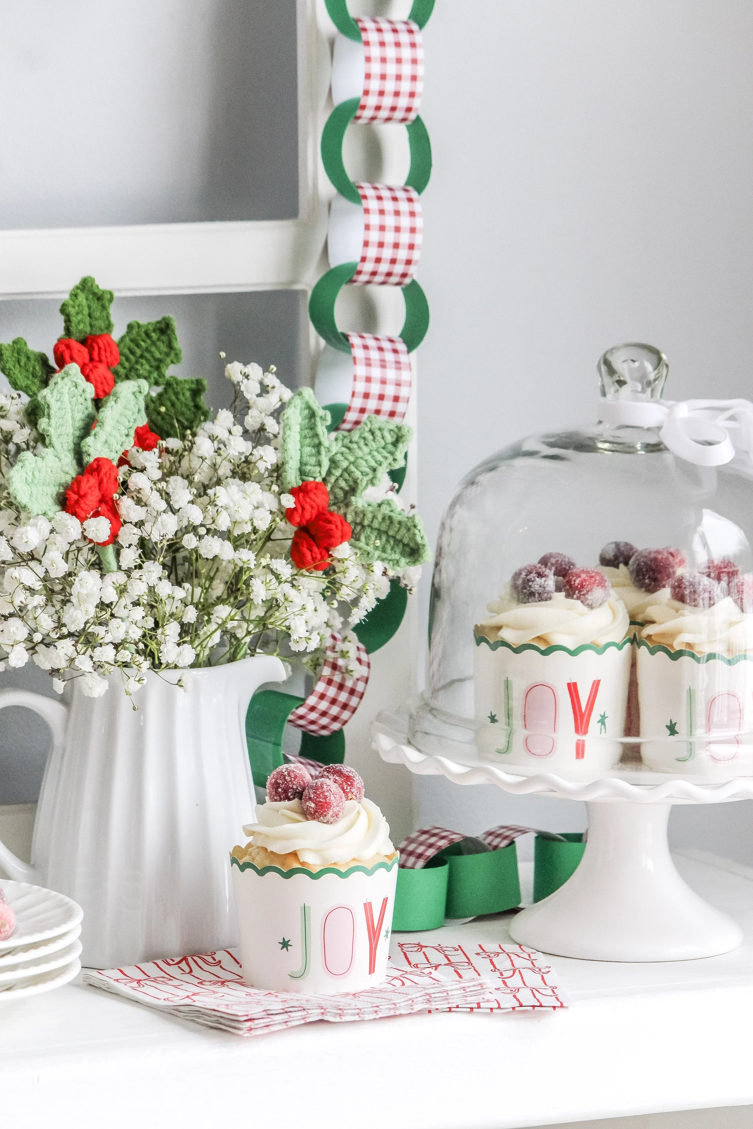 Cupcakes with whipped cream and sugar-coated berries under a glass dome on a white cake stand, a vase with white and red flowers, and holiday decorations with green and red rings and a checkered ribbon.