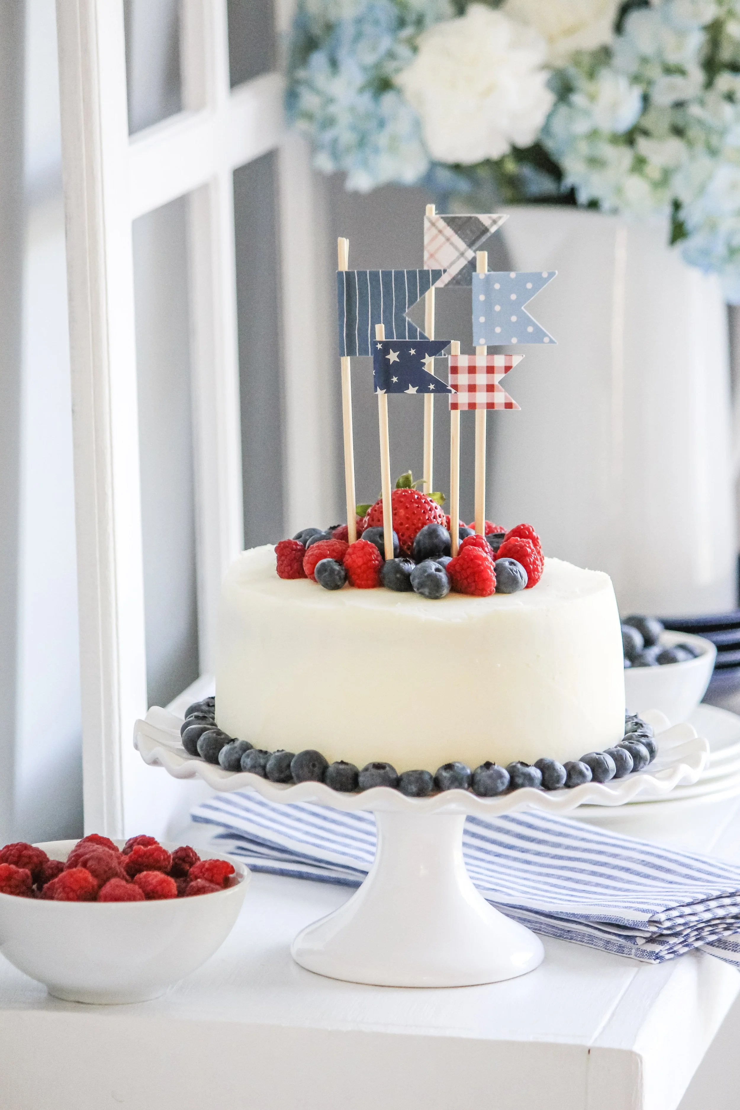 A white frosted cake decorated with fresh blueberries and raspberries, topped with small patriotic flags, on a white cake stand. A bowl of raspberries and blueberries and blue and white striped napkins are nearby.