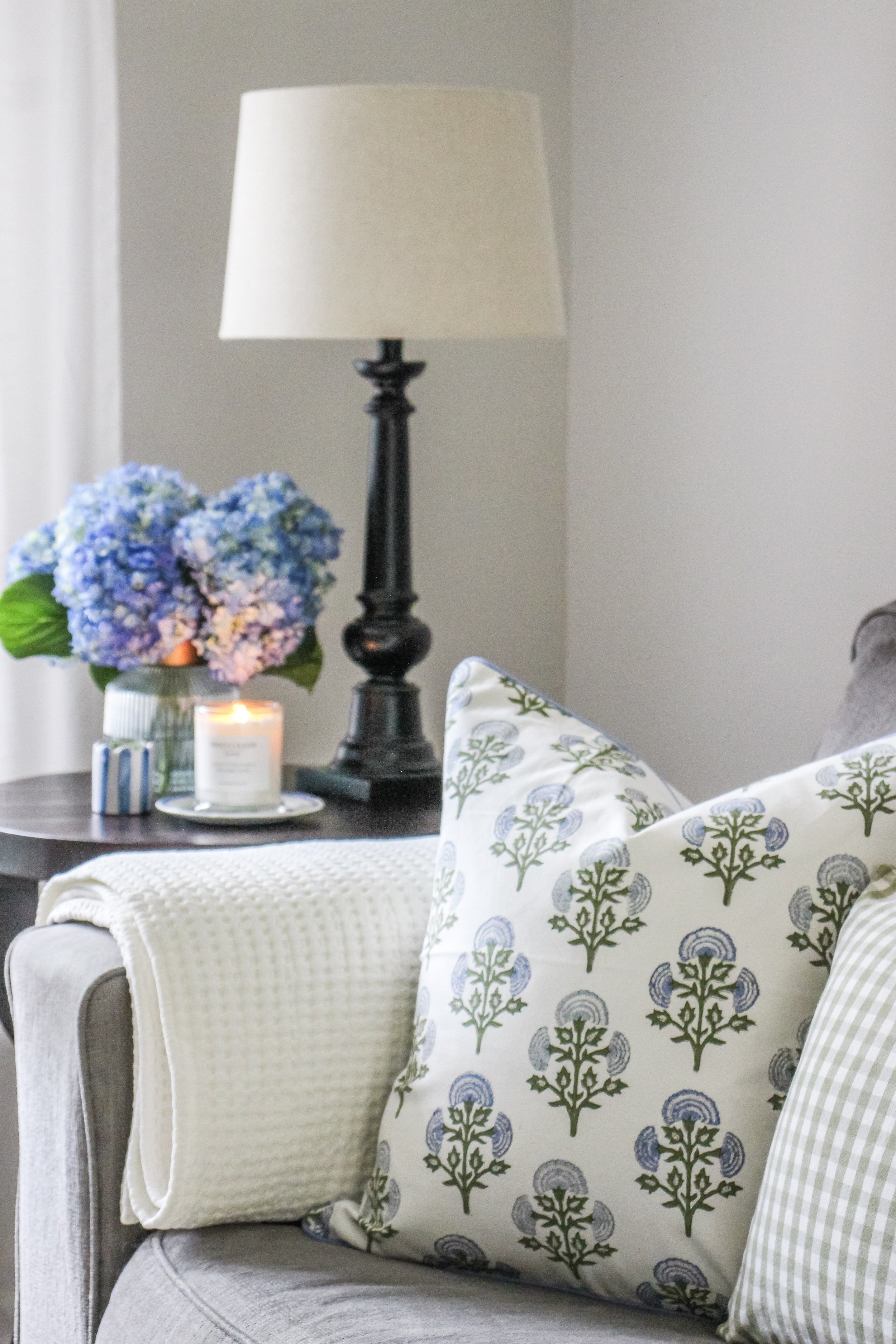 A cozy living room corner with a beige couch, decorative pillows featuring blue flowers and green leaves, a side table with a vase of blue hydrangeas, a lit candle, and a black table lamp with a cream lampshade.