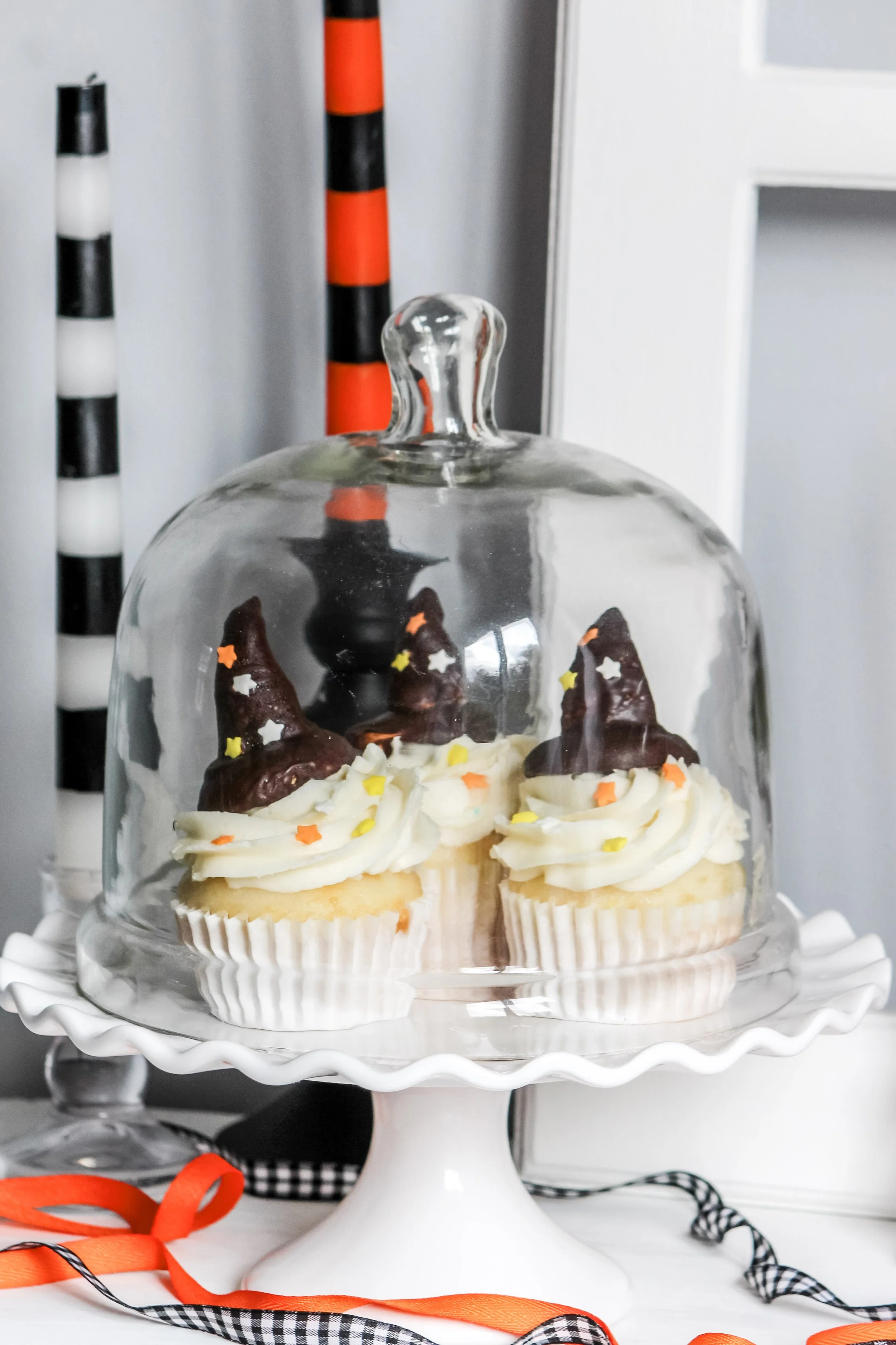 Cupcakes decorated as witches' hats with chocolate and star-shaped sprinkles on a white cake stand with glass cover.