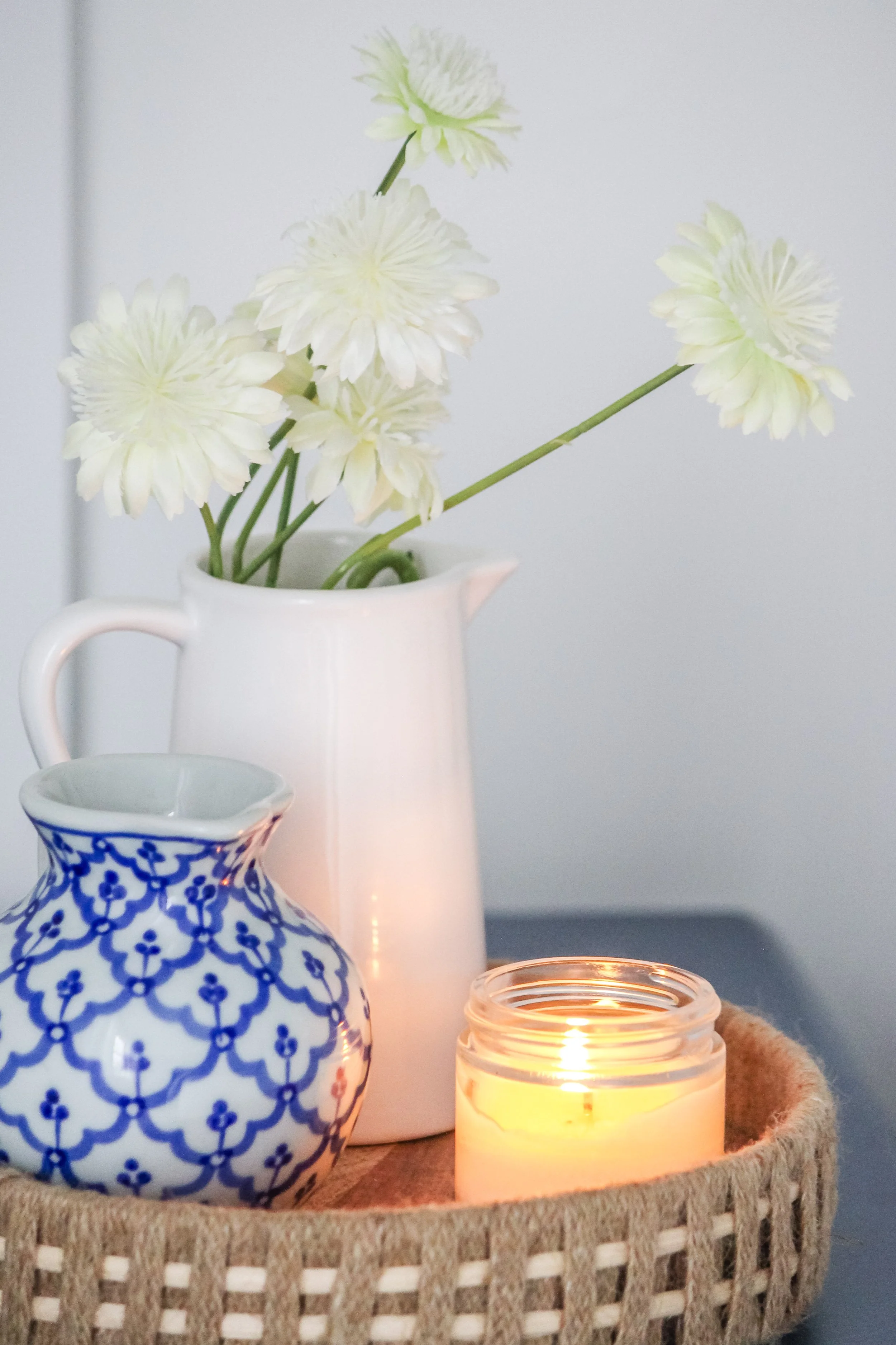 White flower vase with white flowers, a small blue and white patterned vase, and a lit candle in a glass jar on a woven tray.
