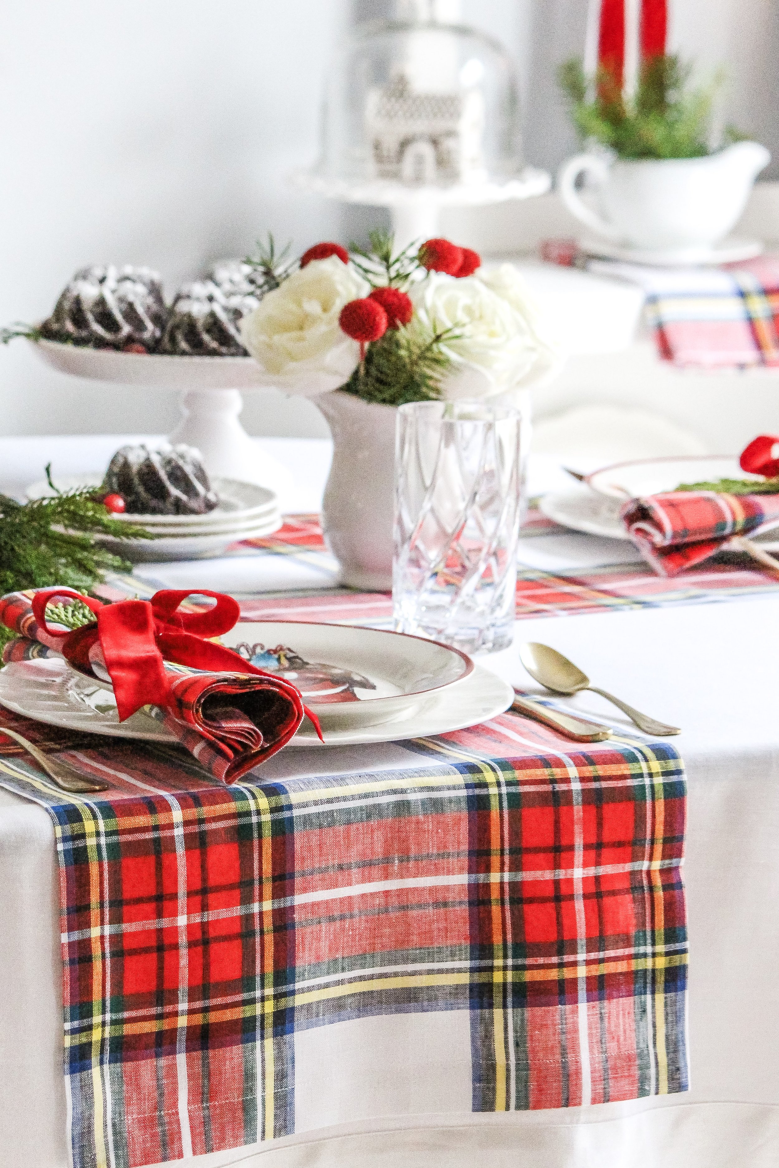 A festive holiday table setting with a red plaid tablecloth, white plates, gold utensils, and clear glassware. It features a centerpiece with red berries, white roses, and greenery, as well as a plate of decorated cookies in the background.