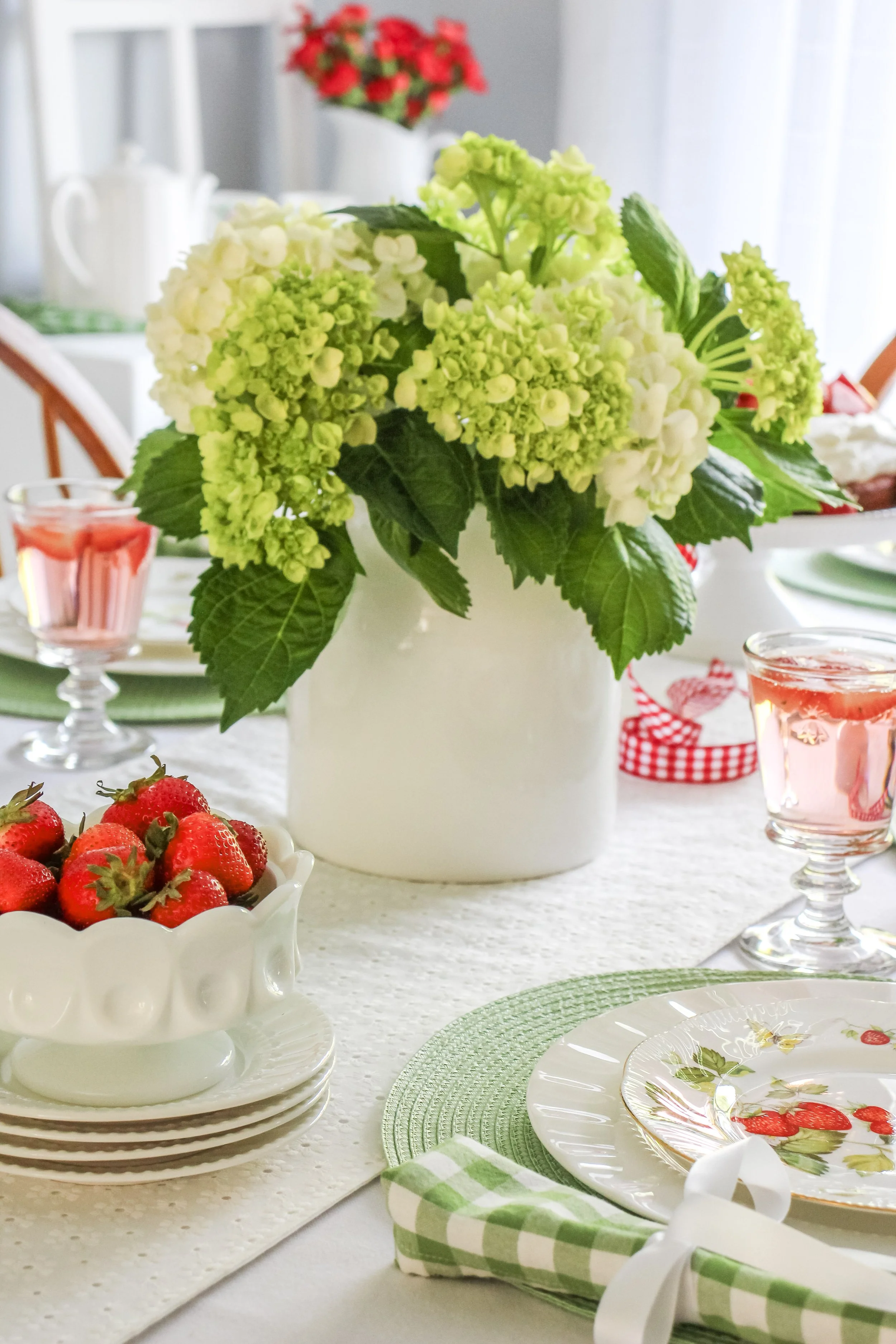 A table setting with a large white vase of green and white hydrangeas as the centerpiece, a bowl of fresh strawberries, and pink drinks in clear glasses, with green and white checkered napkins and strawberry-patterned plates.