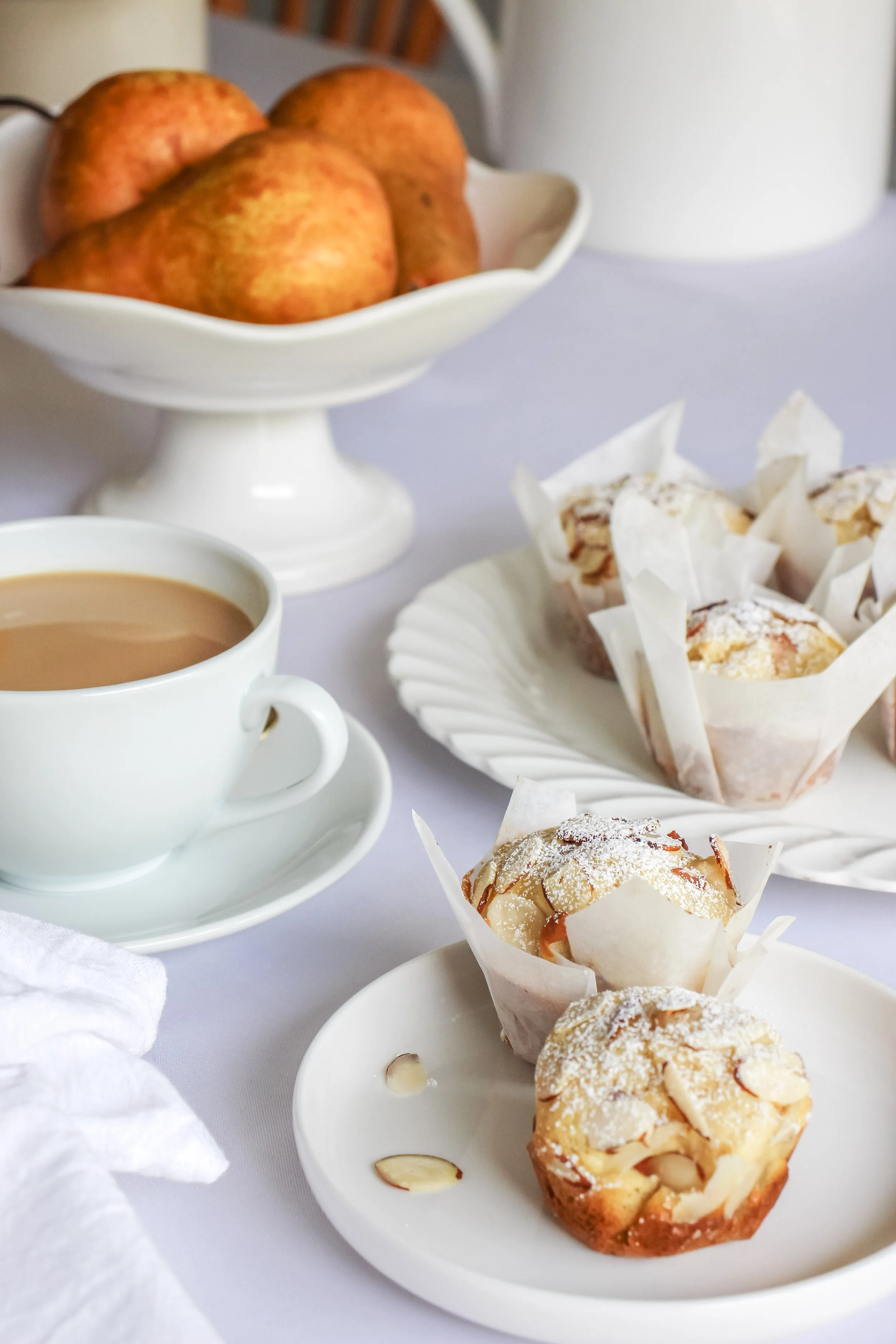 A white plate with two muffins, one topped with powdered sugar and almond slices, on a white tablecloth. In the background, a cup of coffee, a dish of baked apples, and a white bowl filled with apples on a cake stand.