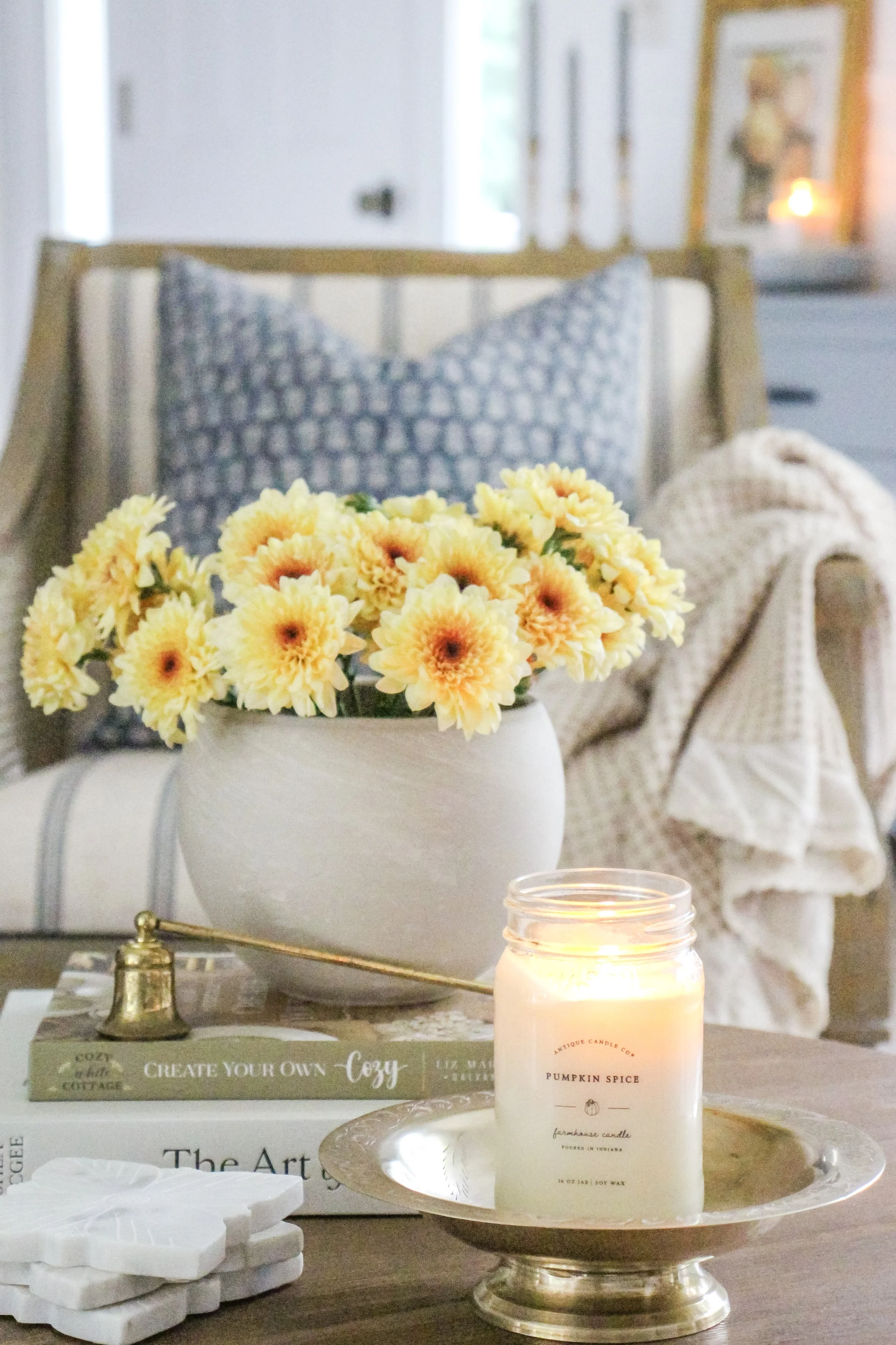 A cozy living room scene with a table holding a lit candle, a white vase of yellow flowers, coasters, and books, with a pillow and blanket on a chair in the background.