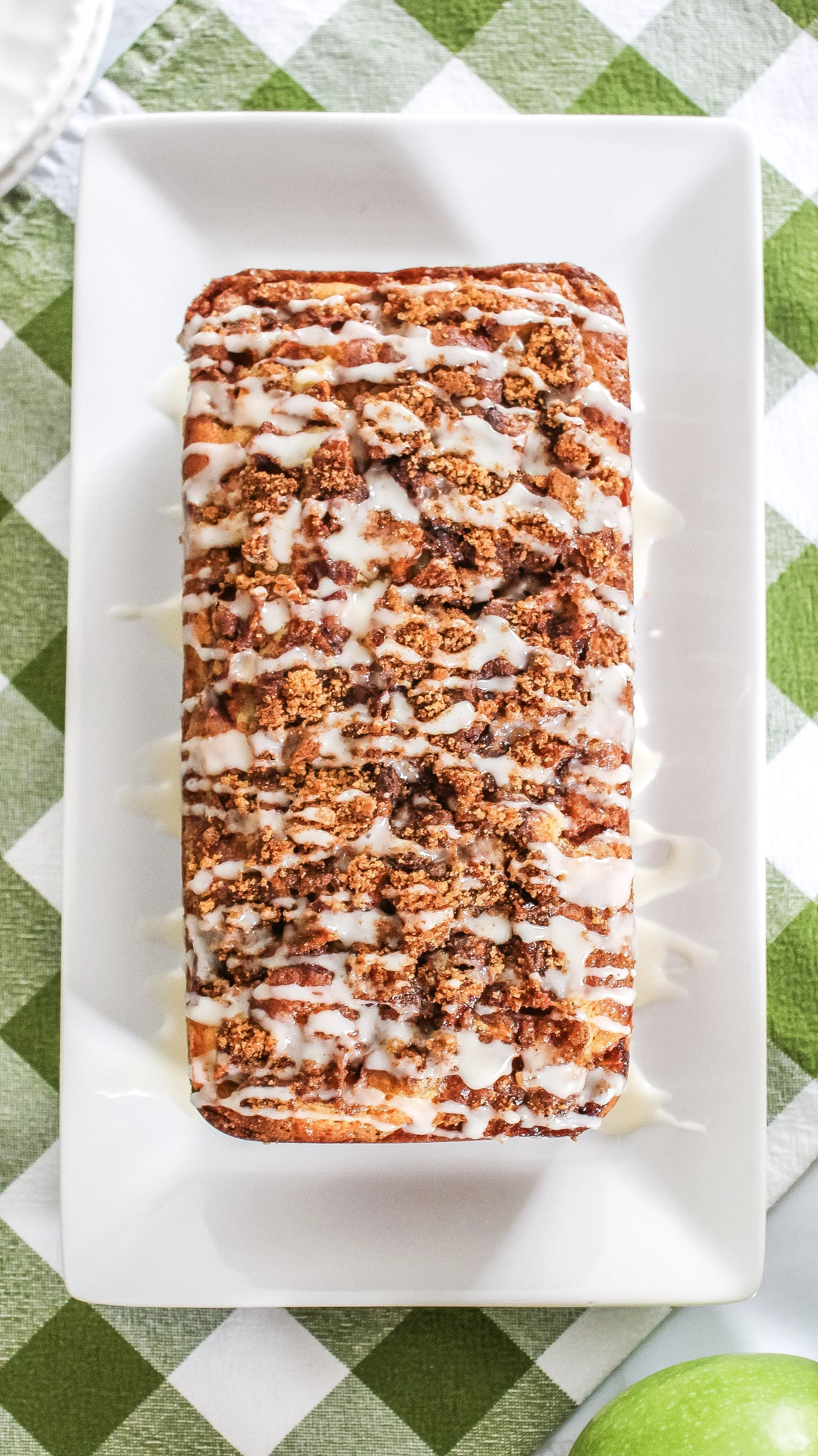 Peach cobbler topped with crumbled streusel and drizzled icing on a white rectangular plate, placed on a green and white checkered tablecloth.