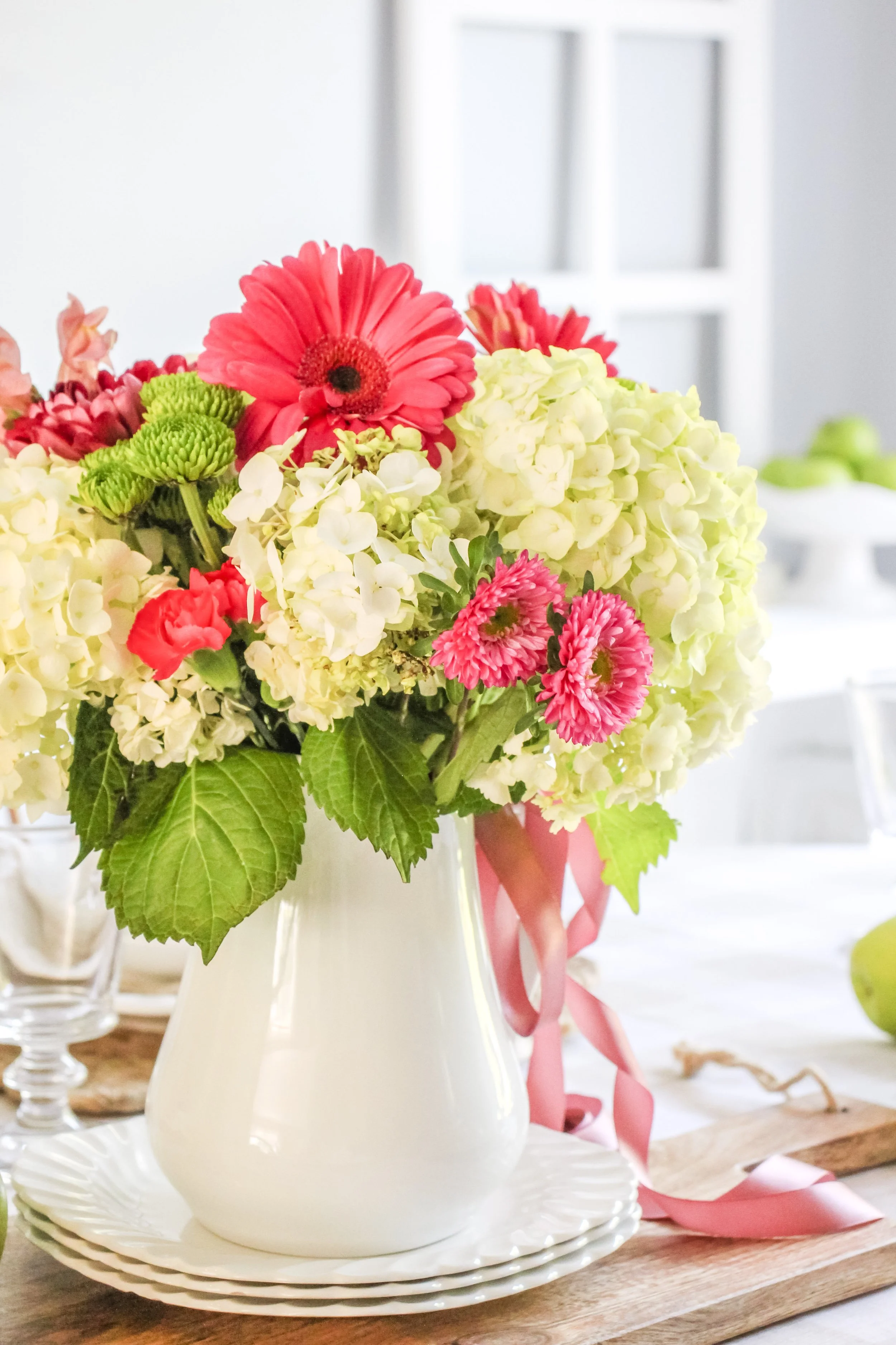 A white ceramic vase filled with pink, white, and green flowers on a wooden table.