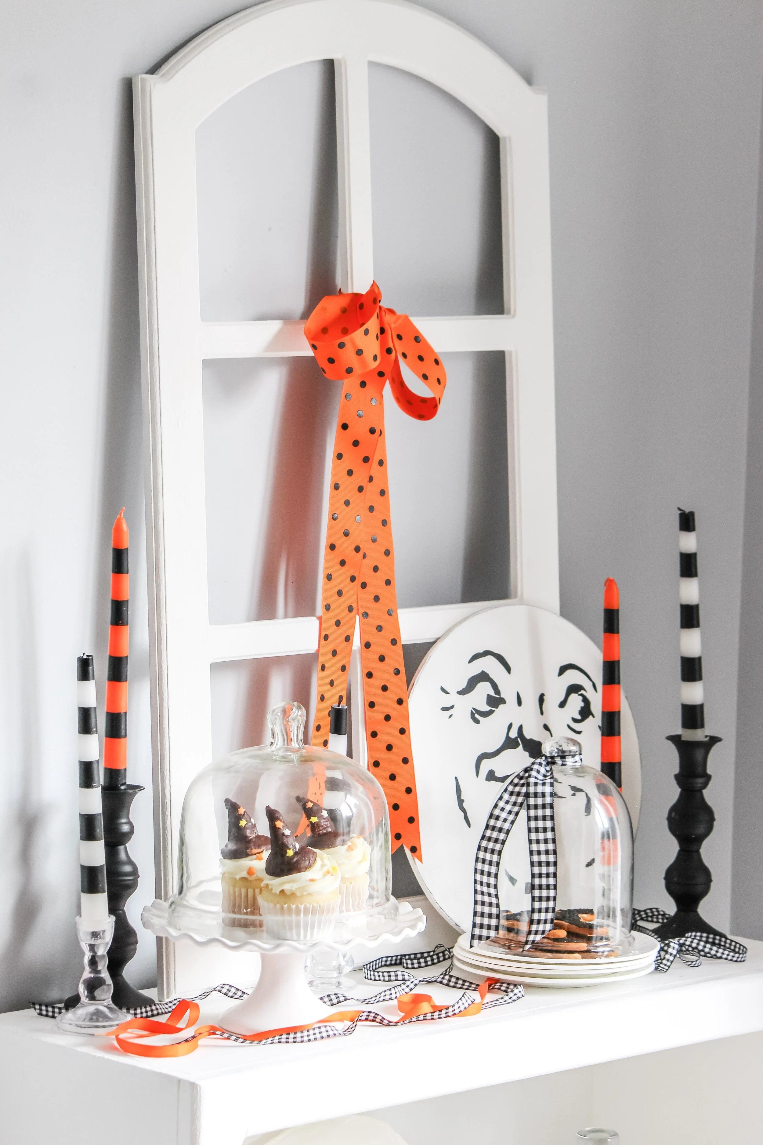 Decorative Halloween display with black, white, and orange candles, cupcakes with witch hat toppers, a black and white checkered ribbon, an orange polka-dotted ribbon, a black and white face illustration, and a white window frame backdrop.