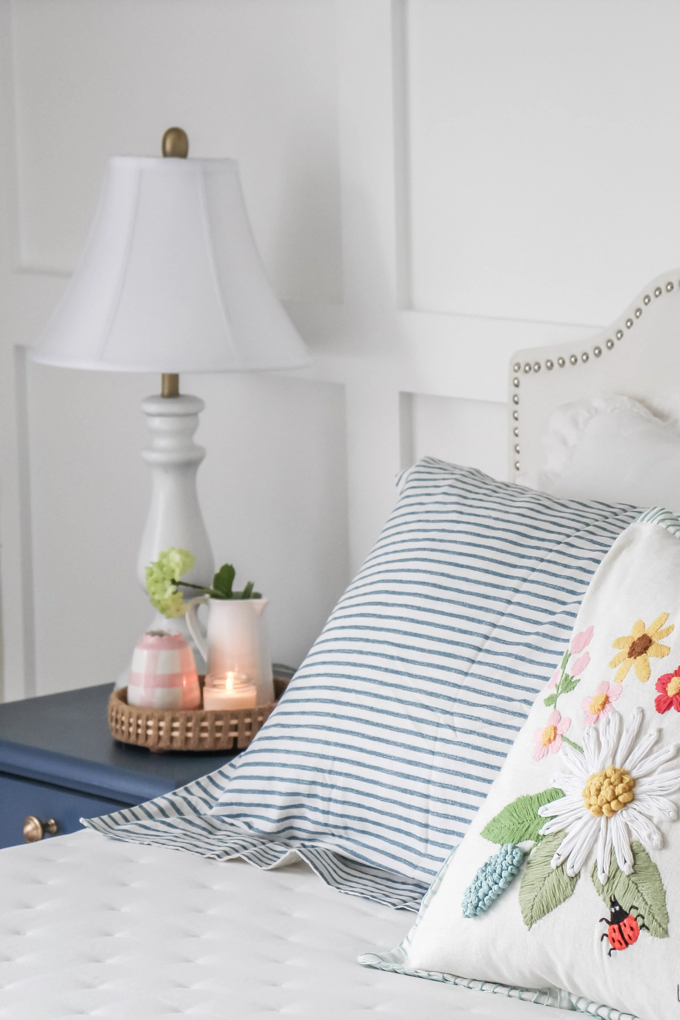 A cozy bedroom with a white headboard, striped pillow, embroidered floral pillow, blue nightstand, a white lamp, a tray with a candle, and a small vase on the nightstand.