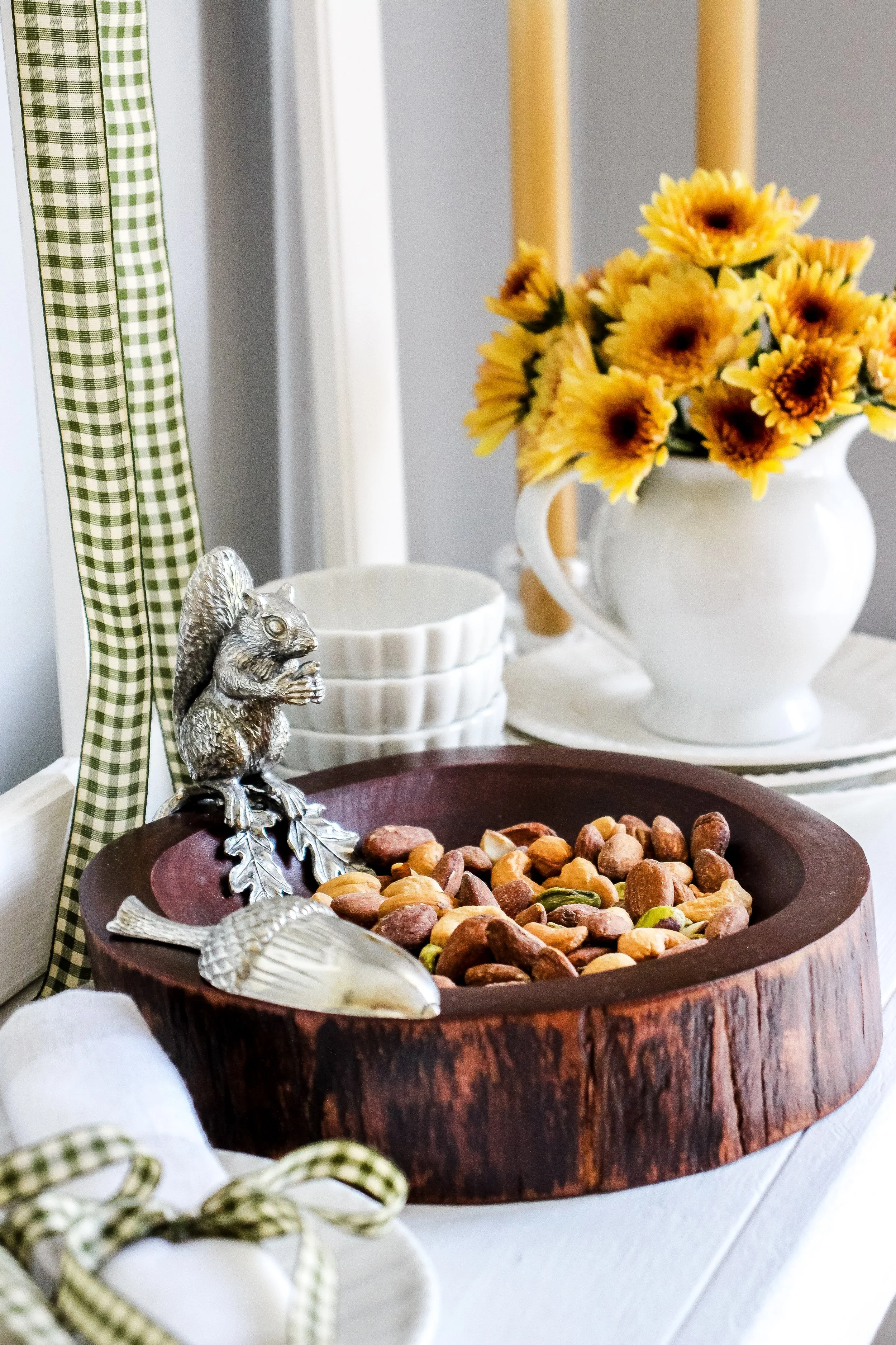Decorative table with a wooden bowl of mixed nuts, a silver squirrel figurine, and a white pitcher filled with yellow sunflowers.