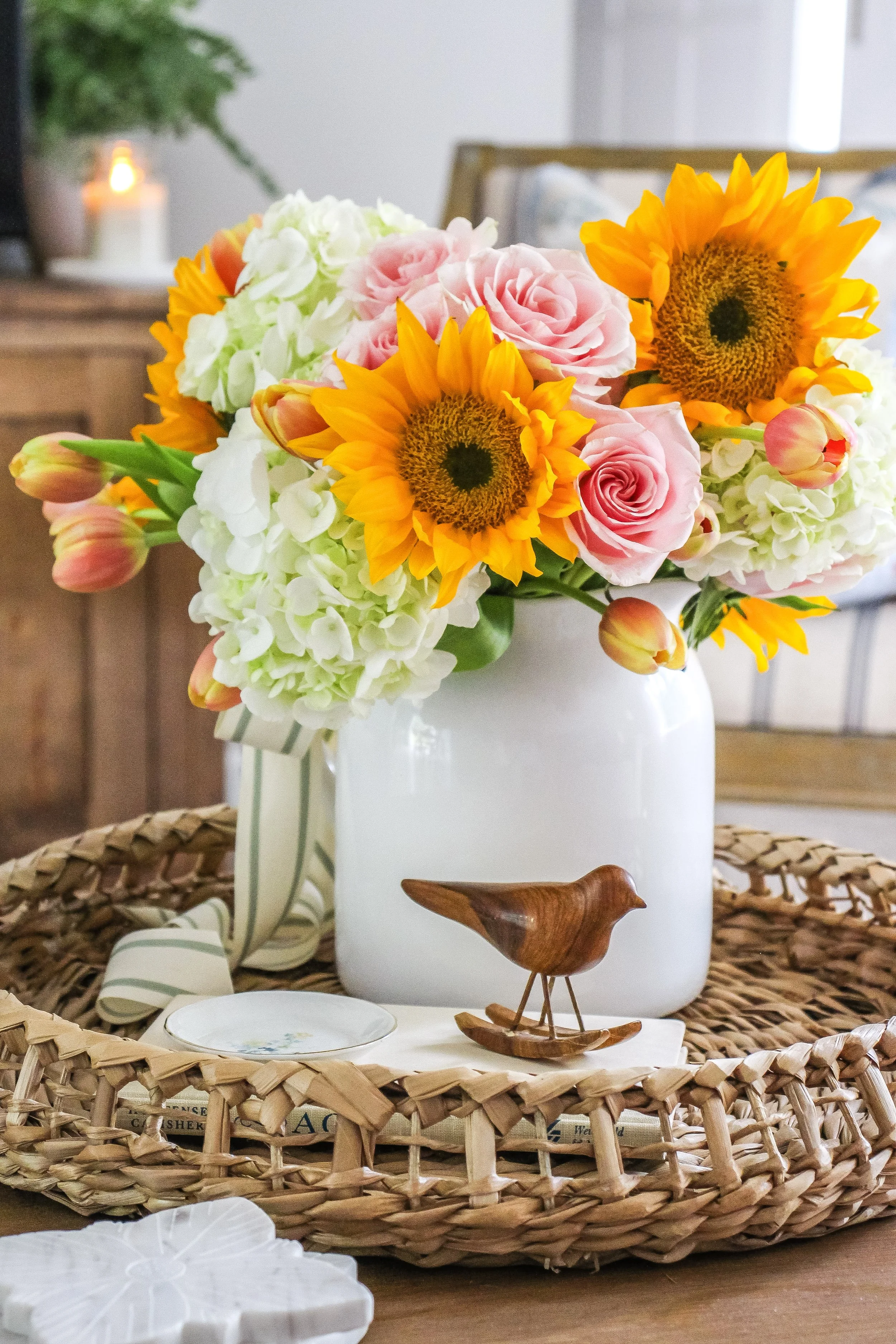 Vase with a bouquet of sunflowers, pink roses, white hydrangeas, and tulips on a wicker tray with decorative bird figurine.