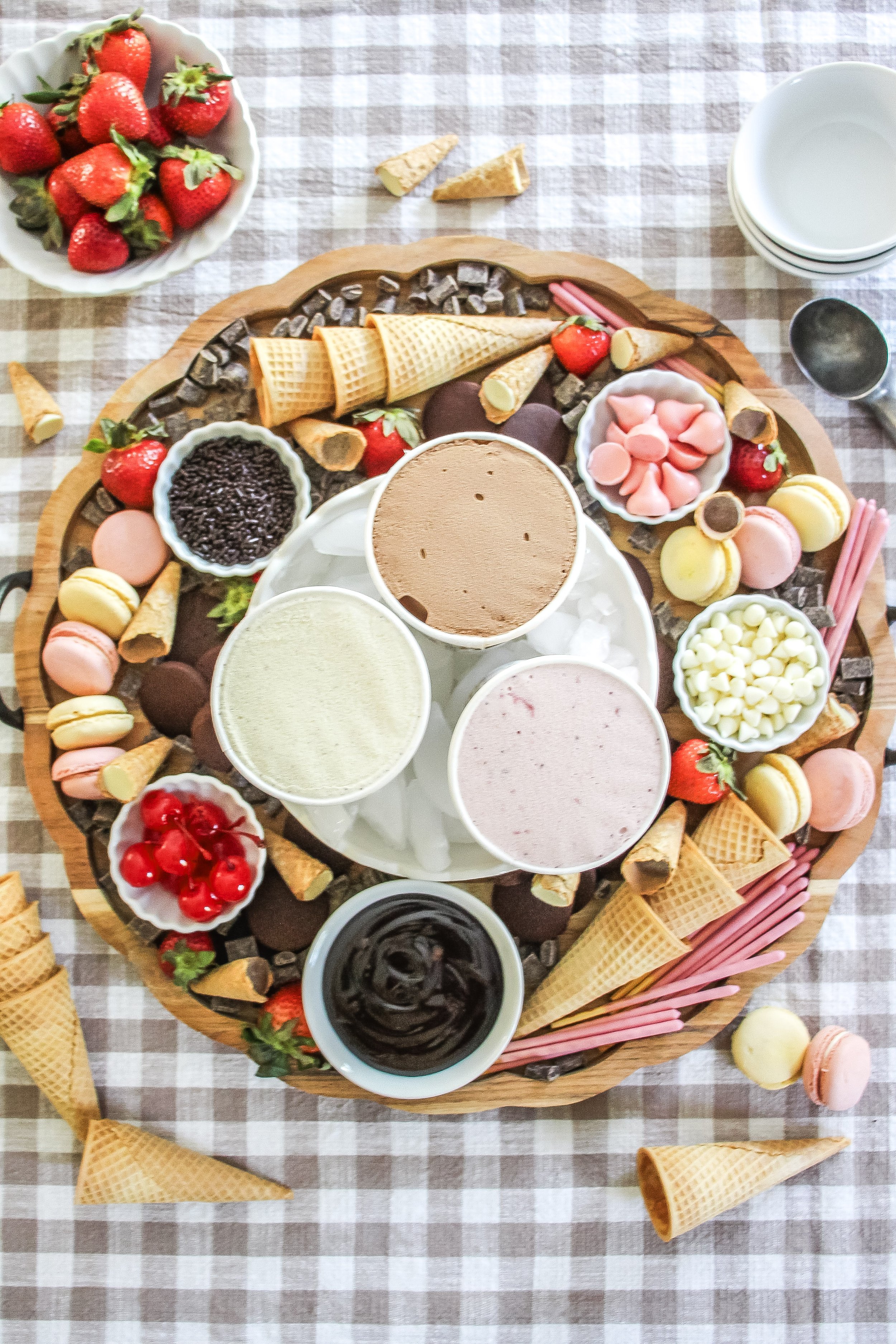 A large wooden platter with assorted ice creams, ice cream cones, gummies, and chocolates, surrounded by strawberries, cherries, and additional ice cream cones on a checkered tablecloth.