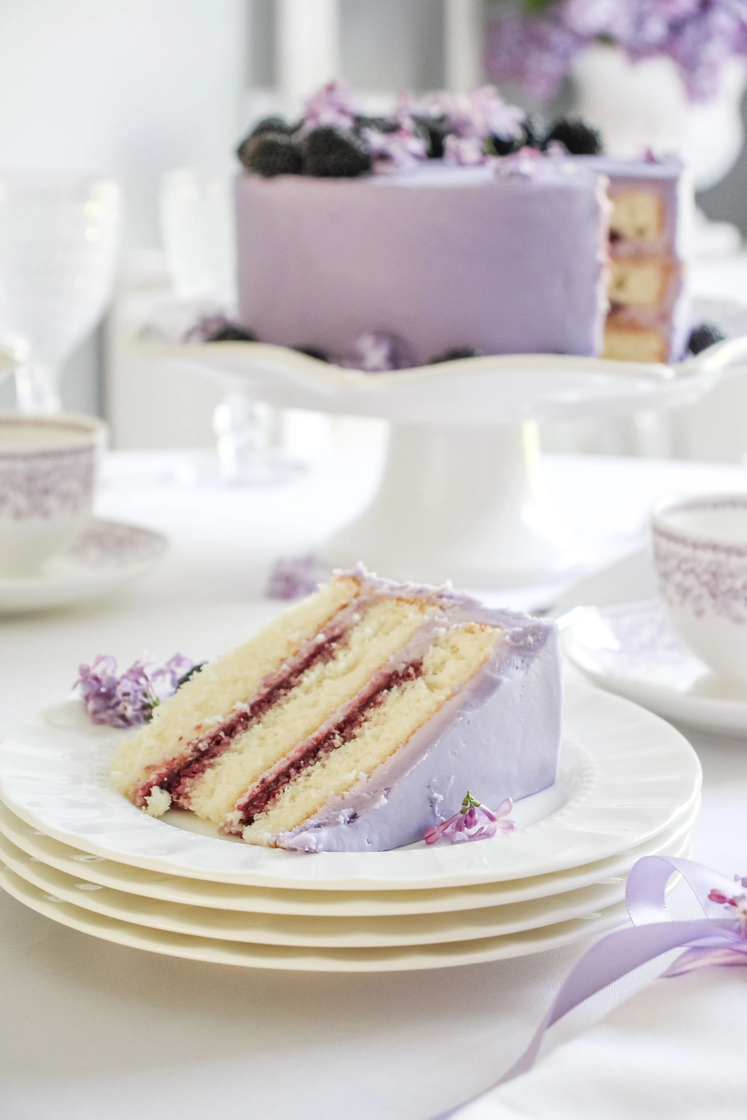 A slice of lavender cake with cream filling on a white plate, with a whole cake in the background decorated with purple frosting and blackberries, set on a white table with matching teacups and saucers.