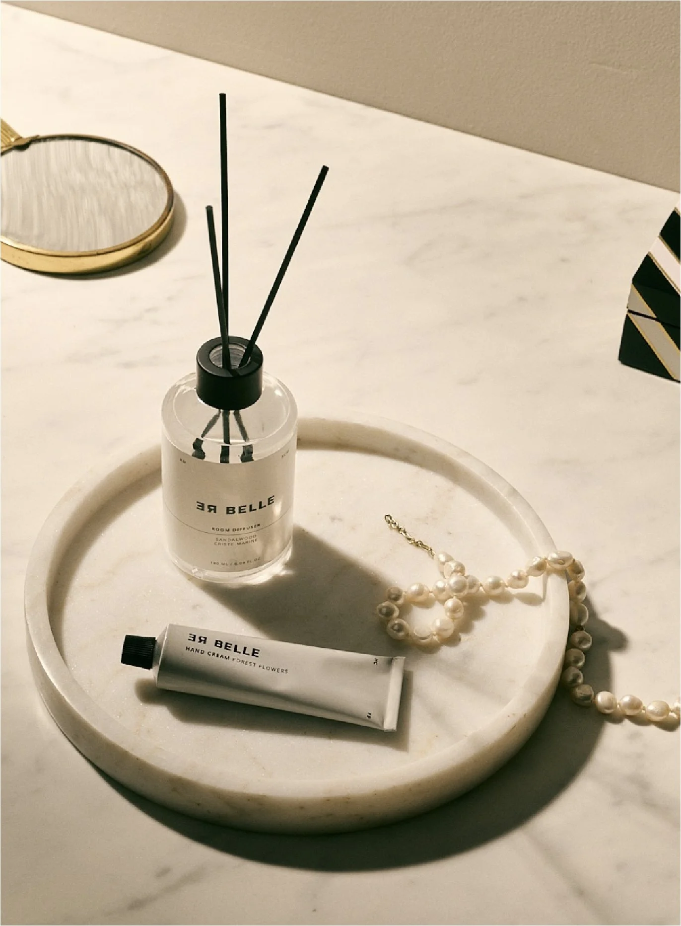 En rund skål med en duftdiffuser, håndcreme og et smykke med perler på en hvid bordplade.