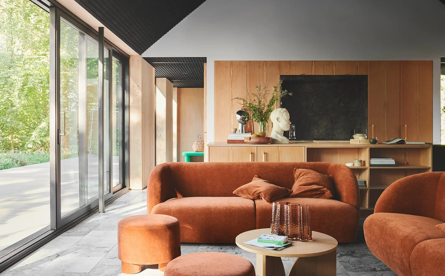 Et moderne stue med store glasdøre, brunt sofa og puf, trækabinettavle med dekorative genstande og plantestil på hylder.